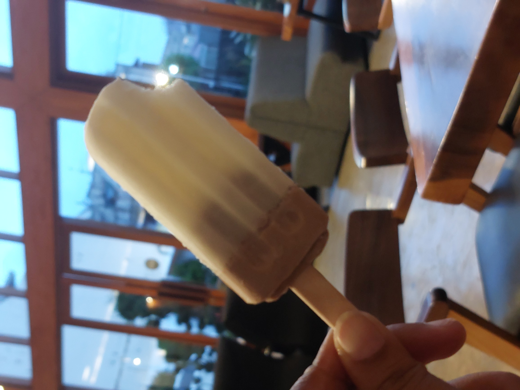 A person is holding a popsicle with a bite taken out of it. The popsicle has a white top and a brown bottom. The person's hand is visible on the left side of the image, holding the wooden stick of the popsicle. The background appears to be an indoor setting with large windows, possibly a cafe or restaurant. Through the windows, some trees and buildings are visible, suggesting an urban or suburban environment. The lighting is dim, indicating it might be evening or an overcast day.