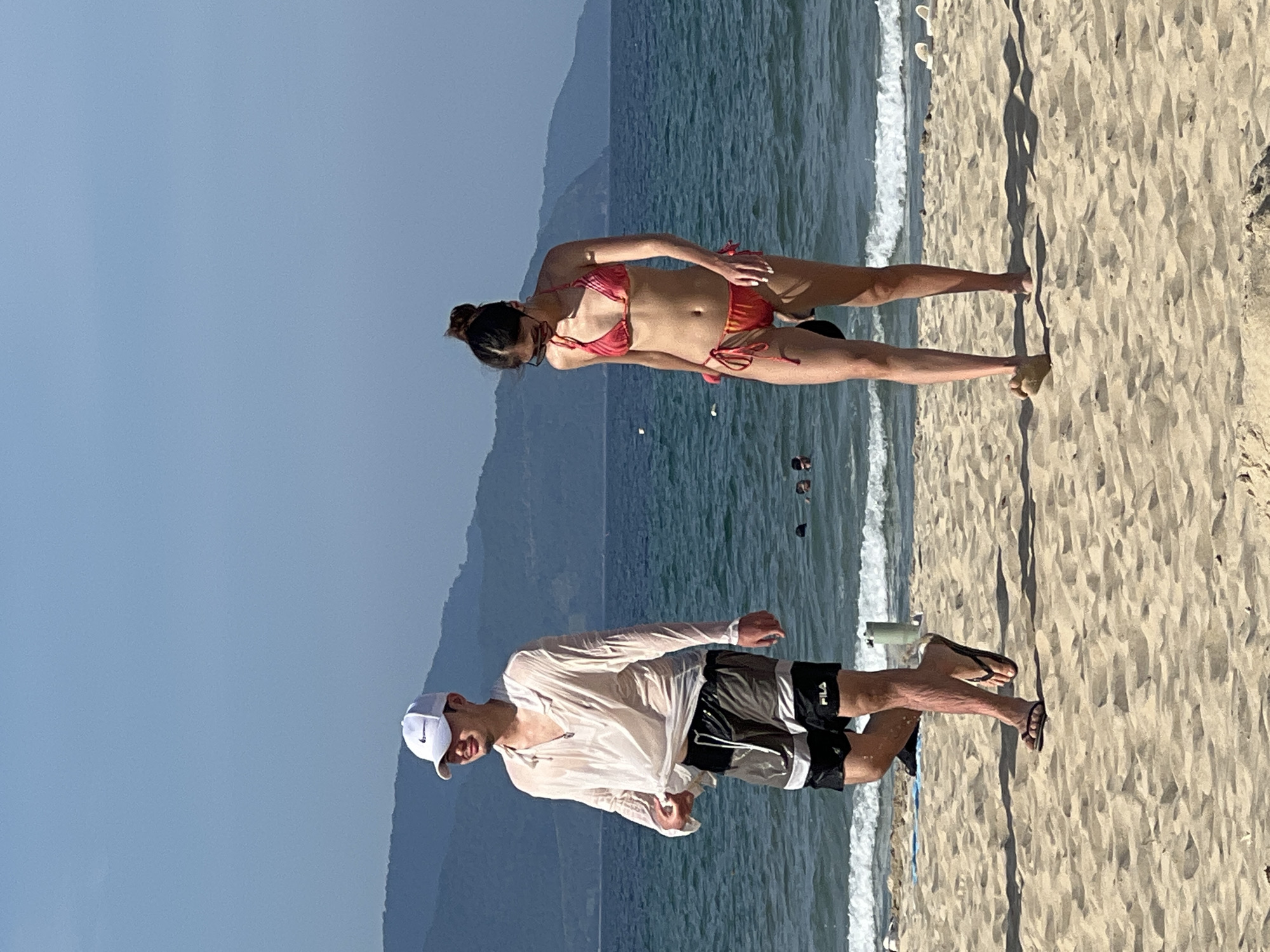 A man and a woman are walking on a sandy beach in Da Nang, Vietnam, with the sea and hills in the background. The man, wearing a white cap, a wet white shirt, and dark swim shorts with a stripe, walks towards the left side of the frame, looking to his left. The woman, in a red and yellow tie-dye bikini and sunglasses, walks towards the right side of the frame, looking down at the sand. The sun is shining brightly, casting shadows on the beach. The ocean has moderate waves, and there are a few other people in the water. The overall scene depicts a casual day at the beach.