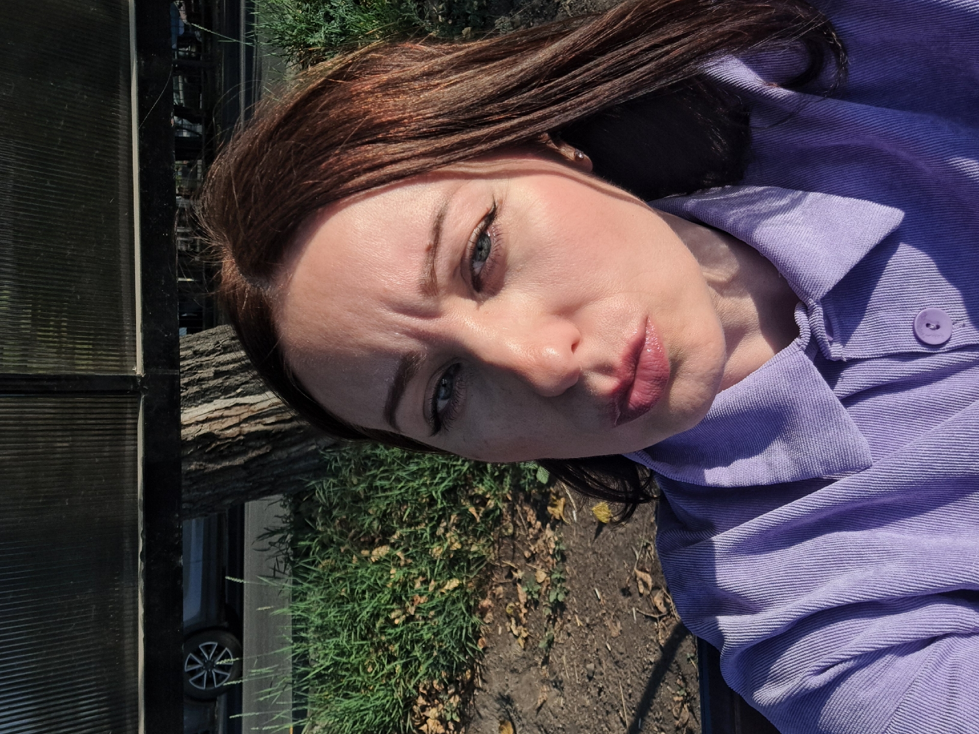 The image is a selfie of a Caucasian woman with reddish-brown hair, taken outdoors during the daytime. She is wearing a light purple corduroy button-up shirt. Her face is angled towards the camera, with her lips slightly pursed. She has makeup on, including eyeliner and blush. The background shows a tree trunk, green grass, dry leaves on the ground, and what appears to be a road with a car in the distance. Above the background is a dark, ribbed structure. The lighting suggests it is sunny, with shadows cast on the woman and the ground. The provided crops confirm these details and offer closer views of the woman's facial features and clothing. The location is identified as Irkutsk, Russia.