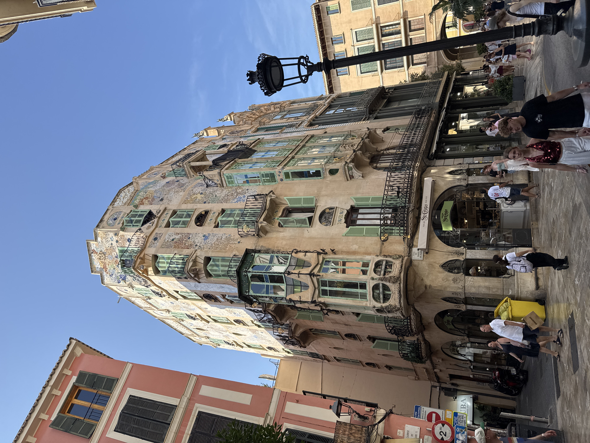 This image captures the facade of the Can Fortuny building, a historic Art Nouveau structure located on the corner of Plaça del Marquès del Palmer and Carrer de Colom in Palma de Mallorca, Spain. The building is characterized by its ornate, multi-colored tiled exterior, featuring intricate patterns and vibrant accents. Several bay windows, some of which are projecting and uniquely shaped, jut out from the facade, adorned with green shutters. Balconies with wrought-iron railings are visible on various levels. The ground floor of the building houses shops, with awnings and large display windows. A lamppost stands in the foreground to the right. The street is paved, and several people are walking and interacting in the scene. The sky is clear and blue.