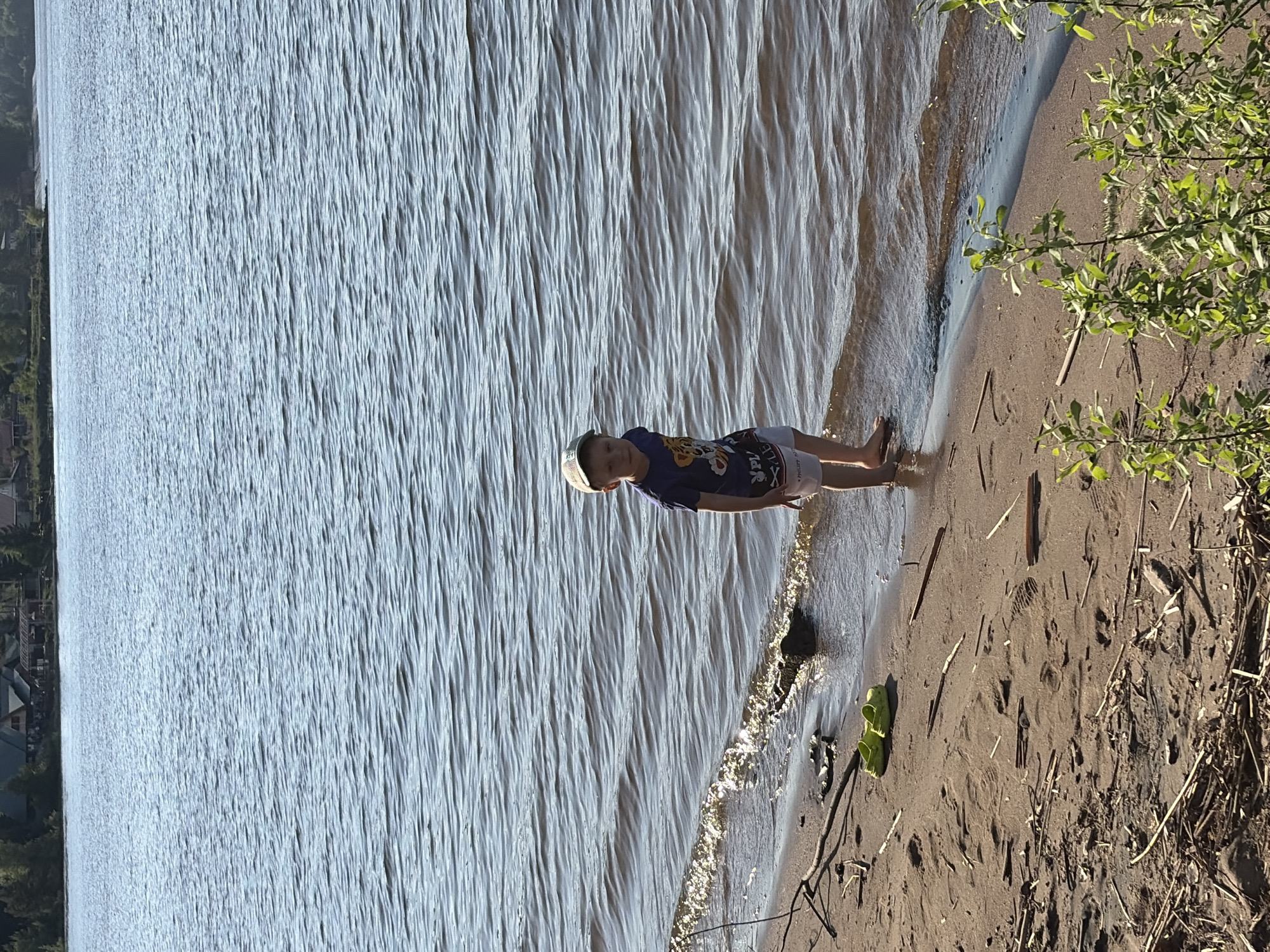 A young boy stands on the shore of a body of water. He wears a dark blue shirt with a cartoon design and white shorts. He has a light-colored hat on his head. The water is light gray with gentle waves. The shoreline is sand. The right side of the image contains greenery. Buildings can be seen across the body of water, indicating this is likely an urban setting. The location is likely Chaikovsky, Russia. The boy is interacting with the water, possibly standing near the edge or wading in it.