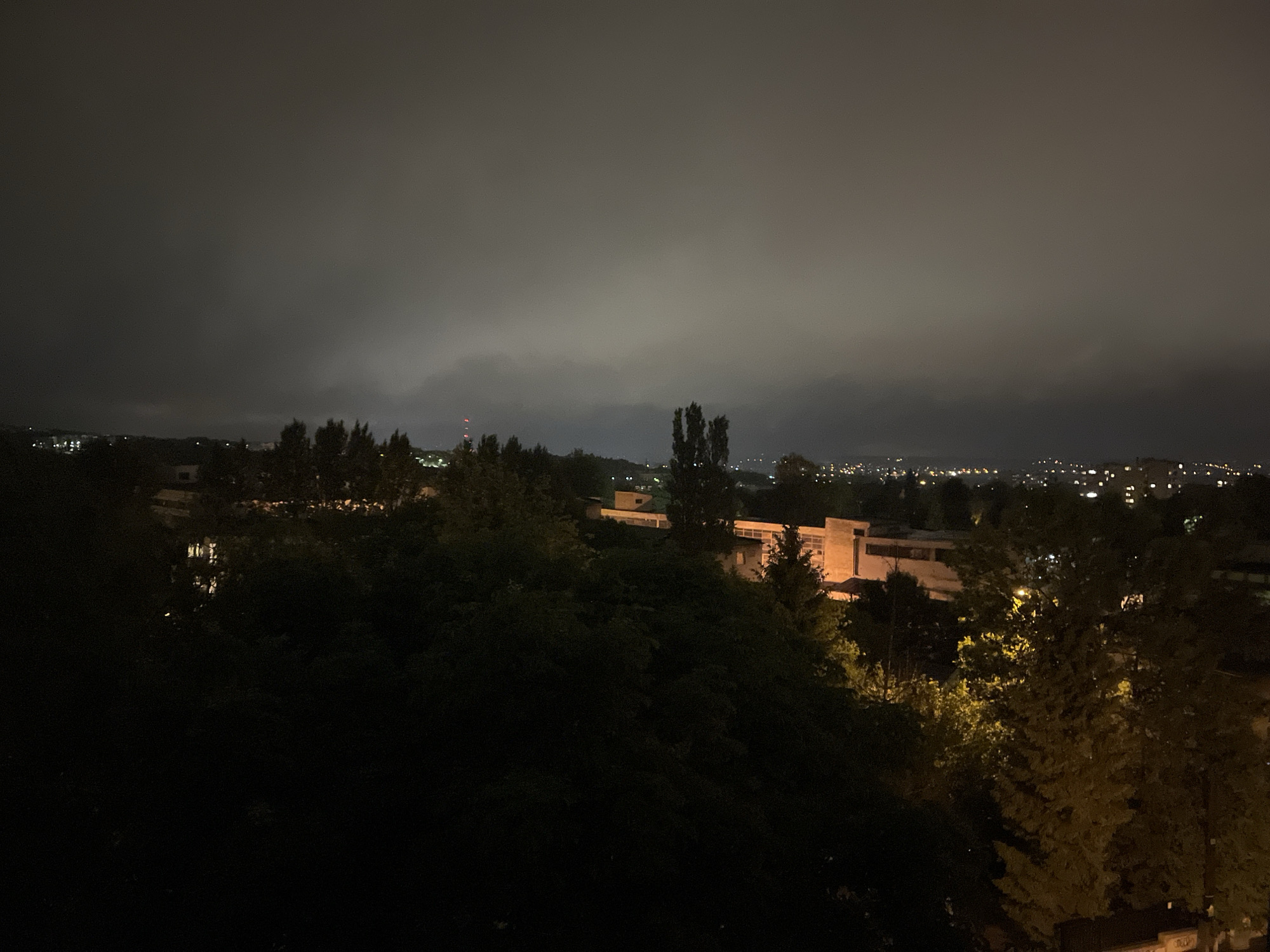 The image presents a panoramic view of a city or town at night, observed from an elevated perspective. The dominant feature is the sky, which is completely overcast with thick, low-hanging clouds that create a uniform, muted gray canopy, reflecting the city lights below.
In the foreground, dense foliage and the dark silhouettes of numerous trees occupy the lower portion of the frame, obscuring the immediate terrain. Within the midground, partially visible through and above the trees, are several buildings illuminated by artificial light. One notable structure on the right-center has a modern, multi-level design with visible windows. A single streetlight on the right casts a warm, yellowish glow on the leaves of nearby trees.
The background reveals the distant cityscape as a scattering of numerous small, bright points of artificial light, stretching across the horizon. A faint red light, possibly an antenna or tower, is barely discernible against the dark clouds on the far left horizon. The overall scene is dimly lit by the urban glow diffused through the heavy cloud cover. There are no visible people or discernible text. The atmosphere is quiet and nocturnal, characterized by the extensive cloudiness.