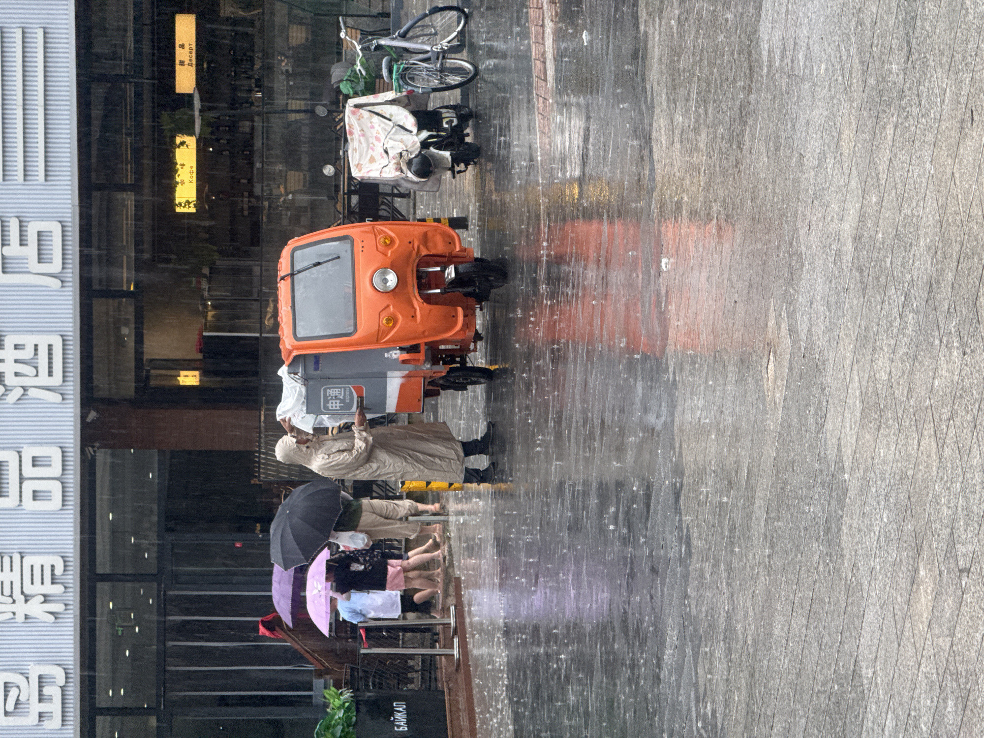 The image depicts a street scene in the city of Qinhuangdao, China, during a rain shower. A person wearing a beige raincoat stands next to an orange three-wheeled vehicle, possibly a delivery vehicle, which has a "申通" logo on its side. In the background, several people are visible holding umbrellas, with purple and black umbrellas being the most prominent. The ground is wet and reflective due to the rain, with visible raindrops impacting the surface. A bicycle and another covered two-wheeled vehicle are parked nearby. The backdrop includes a building with signs in Chinese characters, suggesting a commercial establishment, possibly a hotel or restaurant, as indicated by the characters that translate to "elegant hotel" in the main signage. Smaller signs with what appear to be Russian and Chinese text are also visible, suggesting a diverse clientele or location. The overall atmosphere is that of a common urban setting experiencing inclement weather.