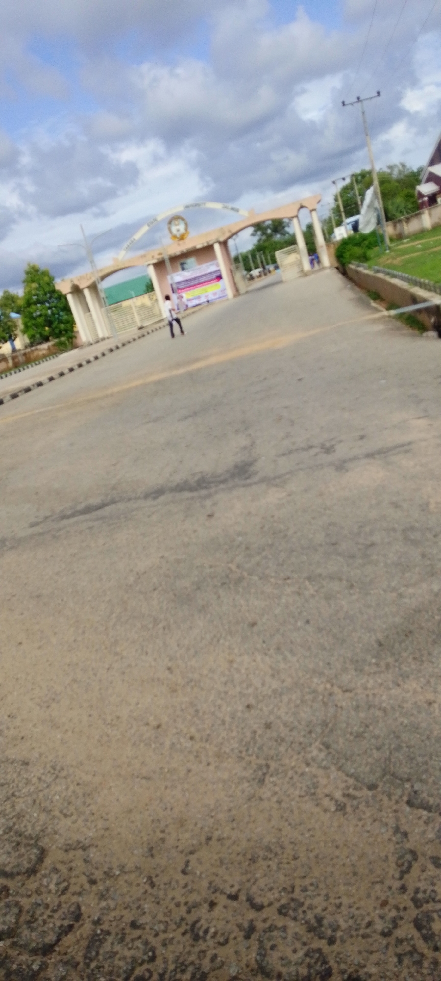 The image depicts the entrance to Taraba State University Jalingo, Nigeria, viewed from the approach road. The foreground consists of a worn asphalt road exhibiting cracks and aggregate exposure, leading towards a prominent, light-colored concrete archway. The archway, identified by the partially legible inscription "TARABA STATE UNIVERSITY JALINGO" and a central emblem, serves as the main entrance. Below the archway, a gatehouse structure is visible, adorned with multiple colorful banners or posters.
One individual, dressed in a light-colored top and dark trousers, stands on the left side of the road near the gatehouse, facing generally towards the entrance. Trees and green vegetation are present on both sides of the road and behind the gate structure. A utility pole with power lines is visible on the right side of the road. The sky is heavily overcast with numerous gray and white cloud formations, indicating diffuse lighting conditions. On the right side, a concrete drainage channel or curb borders a grassy area.