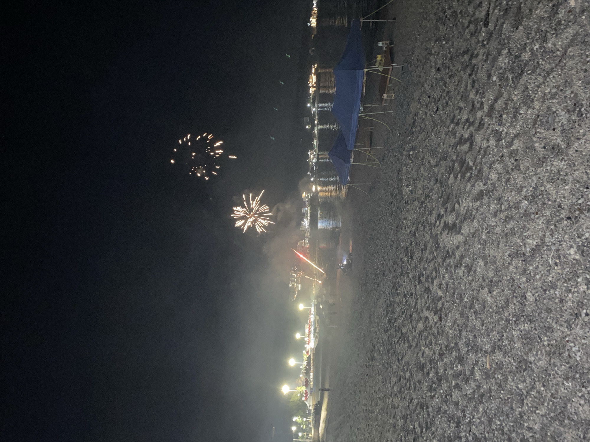 This image captures a nighttime scene on a beach, likely in the city of Gyeongju-si, South Korea, judging by the presence of fireworks. The beach is composed of gravel and sand, with the water of a body of water visible in the background. The sky is dark, illuminated by several bursts of fireworks, some of which are in mid-explosion and others are fading. In the mid-ground, there are several blue canopy tents set up on the beach. Behind the tents, along the shoreline, are numerous bright lights, possibly from streetlights or buildings, and a line of beach umbrellas. A few people are visible on the beach, some near the tents and others further down the shore. A small vehicle, possibly a quad bike, is situated on the gravel near the water's edge. The overall atmosphere suggests a celebratory event, possibly a festival or holiday.