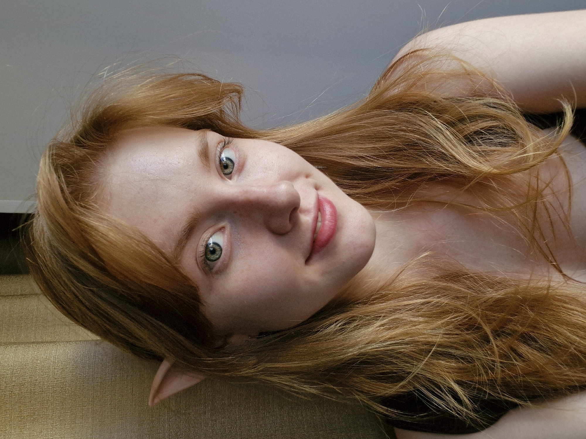 A close-up, tilted shot features a fair-skinned individual with long, red-golden hair and pointed ears. The person has light green eyes and pink lips, with a slight smile. The individual is lying down on a textured surface, possibly fabric. The background is a neutral, light gray. No specific location in Yekaterinburg, Russia, can be determined from the image.