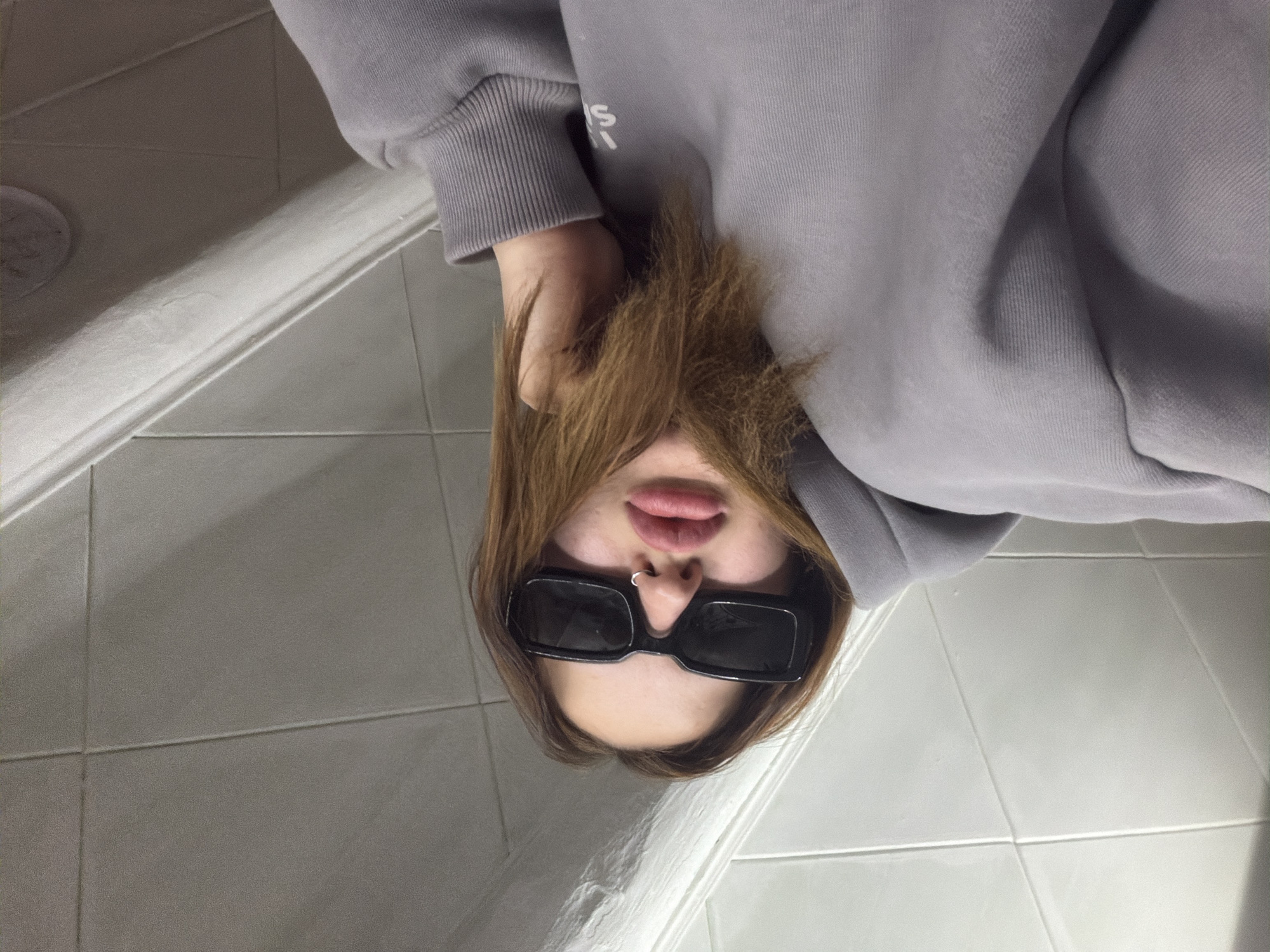 The image displays a close-up, low-angle self-portrait of an individual, likely taken while looking upwards. The subject, whose face is partially obscured, is wearing a light grey hooded sweatshirt with ribbed cuffs. Long, light brown hair falls around their face, and they are wearing black, rectangular sunglasses. A silver septum piercing is visible on the nose, and the lips are slightly parted. White text or a logo, partially visible on the left sleeve or upper torso of the hoodie, appears to start with "IS" or "LS".
The background features light grey or off-white tiled flooring with discernible grout lines. A white, decorative baseboard or molding runs horizontally across the lower part of the scene, suggesting an indoor setting like a bathroom or a utility room. The lighting appears to be artificial and diffused, consistent with an interior space. No specific time of day or outdoor weather conditions can be determined from the image. The overall mood is casual.