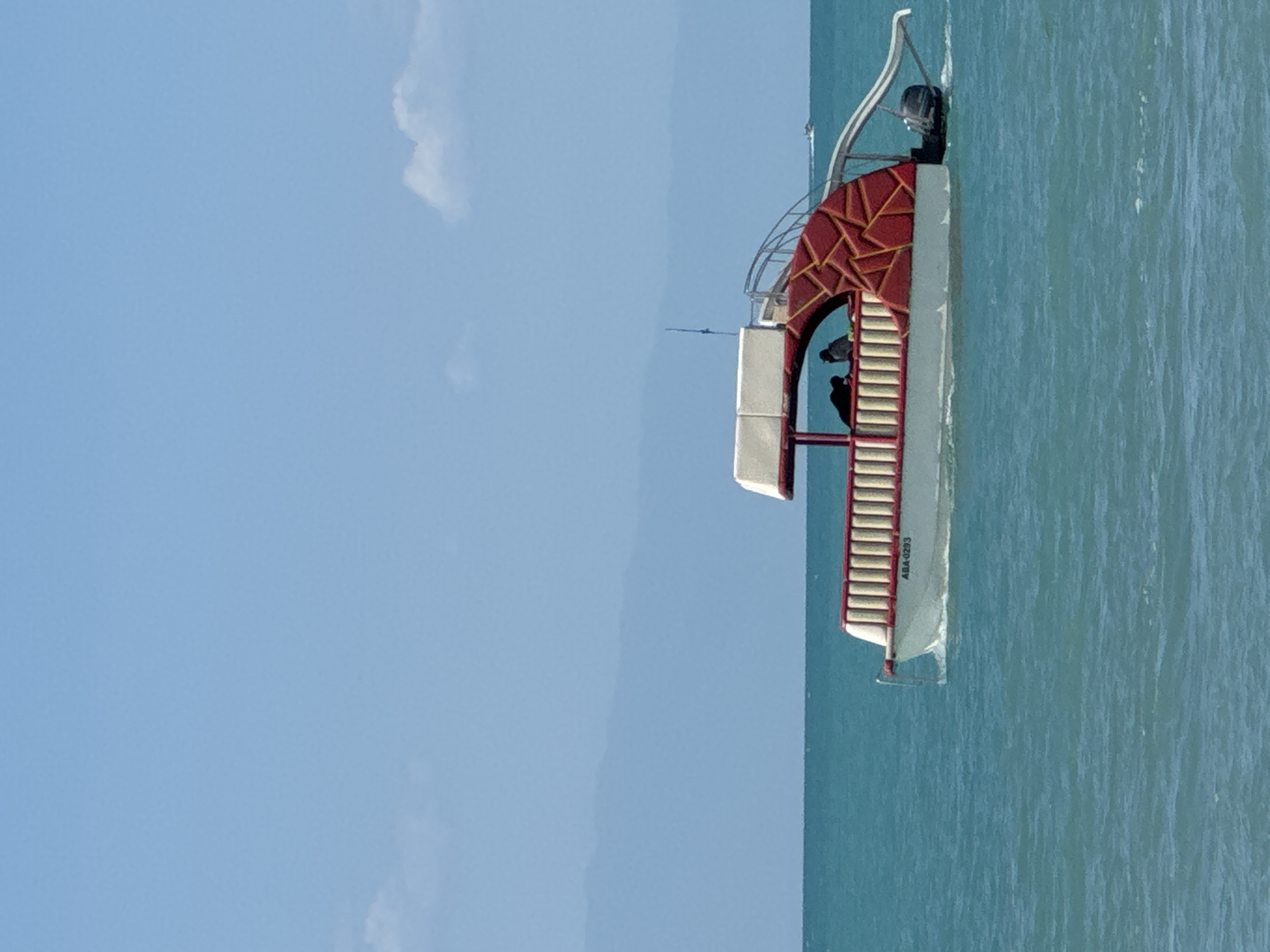 A recreational boat with a slide is on the water under a clear sky with a few scattered clouds. The boat is white with red trim and a patterned red structure on its side. Two individuals are visible on the deck of the boat. The water is turquoise and calm. The image is set in Zharbulak, Kazakhstan.