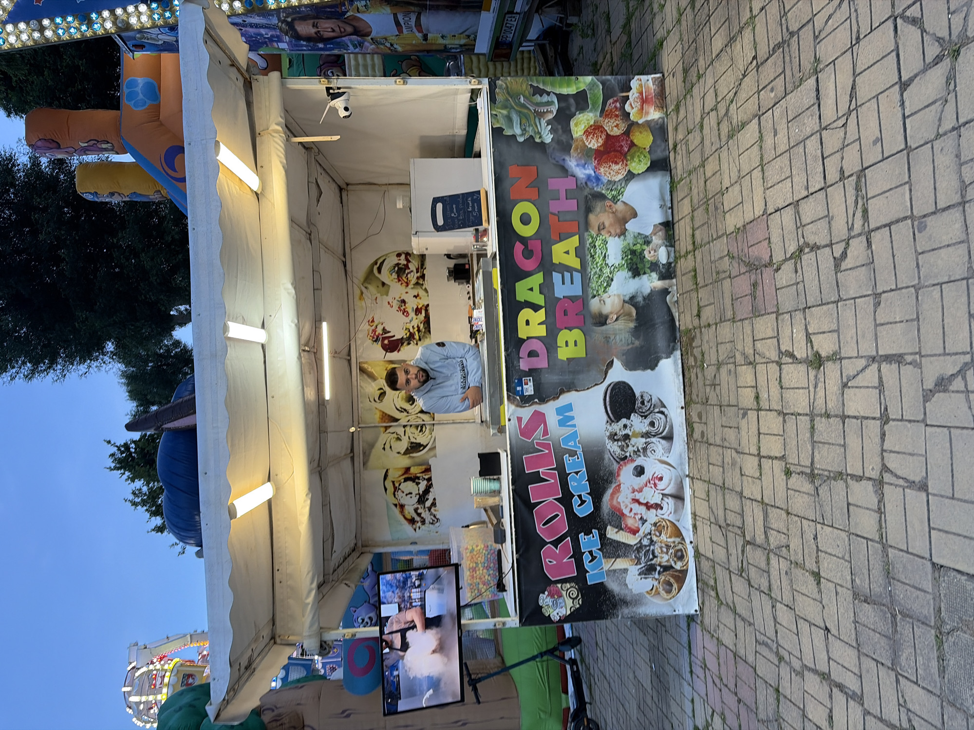 A man stands behind a food cart offering "Rolls Ice Cream" and "Dragon Breath" in an outdoor setting. The cart has a white awning illuminated by fluorescent lights. The front of the cart is decorated with images of ice cream rolls, a dragon, and people blowing smoke. A television screen on the left side of the cart displays a video of ice cream being made. There are also inflatables and amusement park rides visible in the background. The ground is paved with brick-like tiles. The sky is a pale blue, suggesting it is either early morning or late evening.