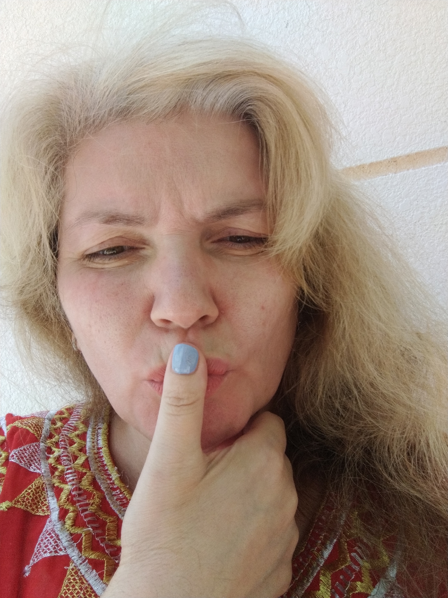 A woman is pictured in a close-up selfie. Her expression is one of contemplation or secrecy, with her index finger touching her lips in a "shhh" gesture. She has fair skin, light blonde hair, and her nails are painted a light blue color. She is wearing a red and gold embroidered top. The background appears to be a plain, light-colored wall. While the image does not contain sufficient information to confirm the specific location in Al Mas'oudi, United Arab Emirates, there are no elements in the image which negate the possibility of this location.