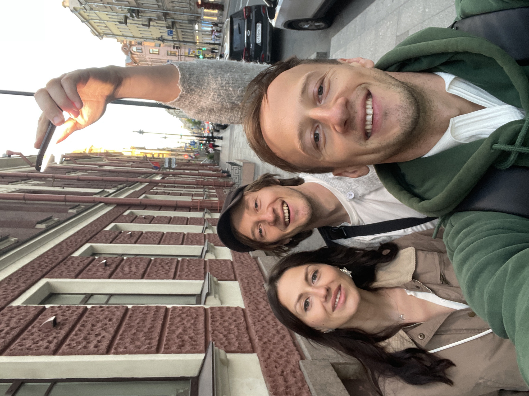 Three people are taking a selfie on a street in Saint Petersburg, Russia, with buildings and a street with traffic in the background. The person in the foreground, on the right, is a man with short brown hair and stubble, wearing a green hoodie over a white collared shirt. To his left is a man with brown hair and a black baseball cap, wearing a light gray sweater. To the far left is a woman with long dark brown hair, wearing a tan jacket over a white top and a necklace with a crescent moon pendant. Her left ear has a star-shaped earring. The person in the background on the right is holding up a small light fixture. The buildings in the background are typical of Saint Petersburg architecture, with tall windows and ornate facades. The street appears to be a busy urban area with cars and pedestrians. The lighting suggests it is late afternoon or early evening.