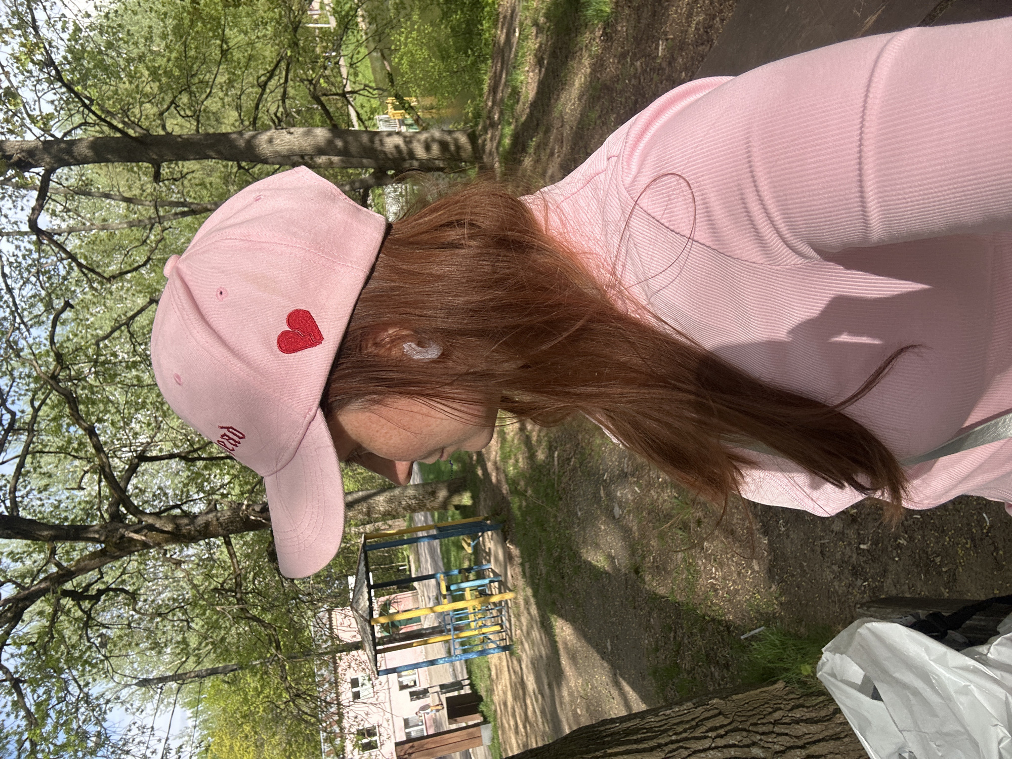 The image shows a person with light brown hair, wearing a pink long-sleeved shirt and a pink baseball cap with a red heart on the side, standing in a park-like setting. The background includes trees with green leaves, a dirt path, and playground equipment painted blue and yellow in the distance. A building is visible in the background. Based on these visual cues, it is plausible that the location could be a park area within the city of Nizhny Novgorod, Russia.