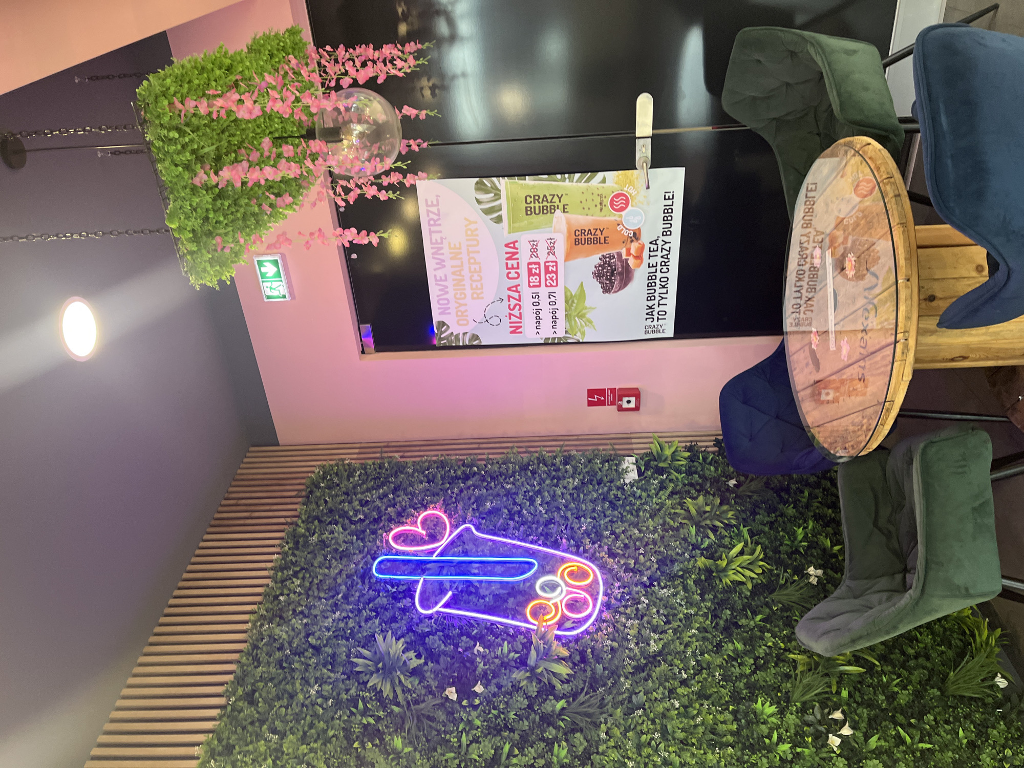 The image shows an interior scene of a bubble tea shop. In the foreground, a round wooden table with a glass top is surrounded by dark teal chairs. On the wall to the left, a large, vibrant neon sign in the shape of a bubble tea cup with a heart and straw glows in neon pink and blue against a backdrop of artificial greenery. To the right of the neon sign, a red fire alarm and extinguisher box is mounted on the wall. Further to the right, a dark door is visible, with a poster displayed on it featuring images of bubble tea drinks and text in Polish. Above the door, a decorative hanging plant with pink flowers dangles from the ceiling. The ceiling is dark gray, and the walls are painted in a light pink color.