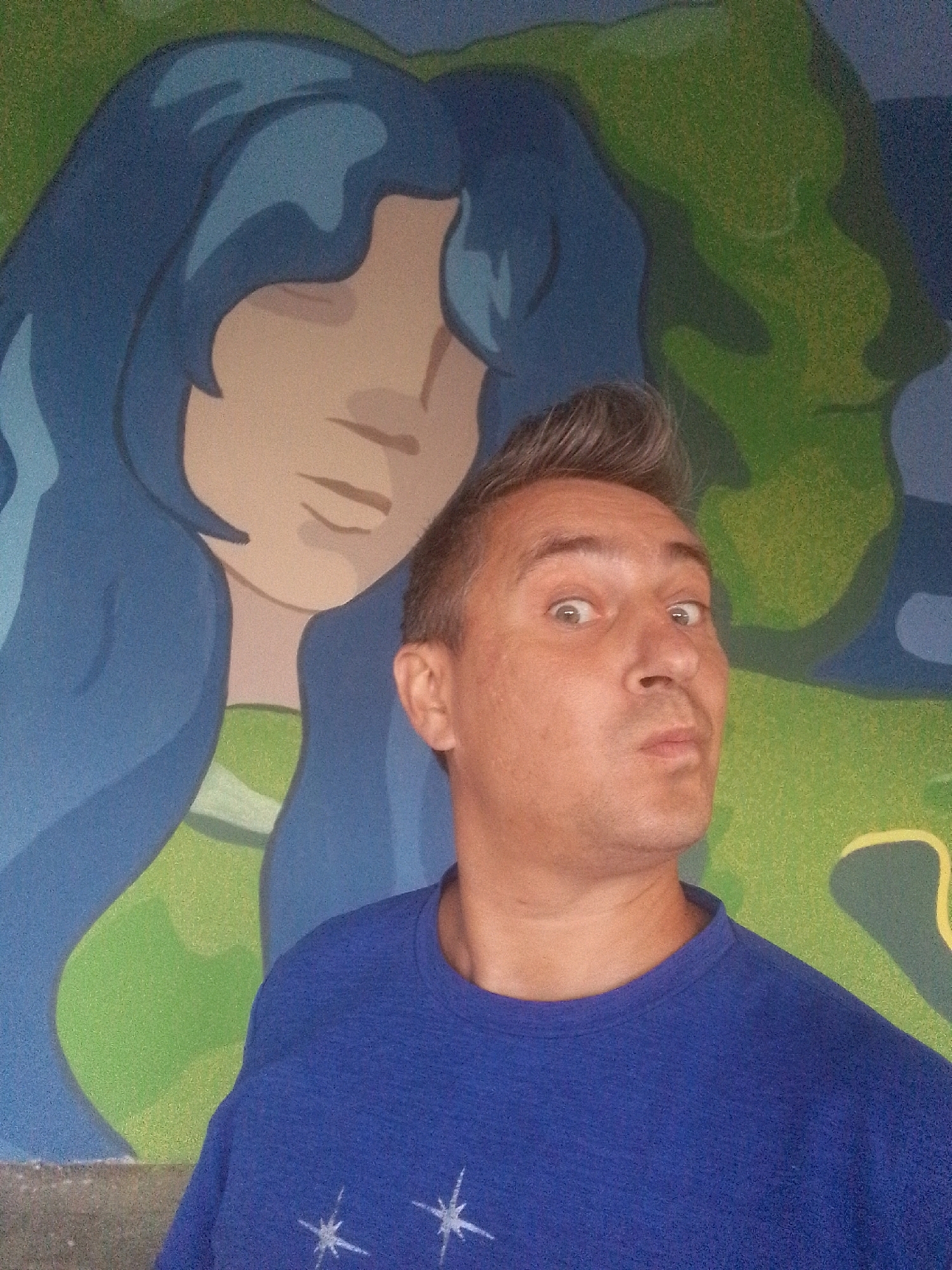 A selfie is taken by a man with short, graying hair styled upward. He is wearing a blue t-shirt with white star-like patterns. The man has a surprised expression with wide eyes and pursed lips. Behind him is a mural with a stylized depiction of a woman with long, blue hair and a green garment. The background of the mural is a mottled green and blue. The man is positioned in the foreground, with his face taking up a significant portion of the frame. The image appears to be taken indoors or in a sheltered outdoor space, given the lighting and the appearance of the mural. The mural itself suggests an artistic or decorative setting, possibly a public space, cafe, or a private residence with a mural. No specific landmarks or identifiable features of Tbilisi, Georgia, are visible in the image.
