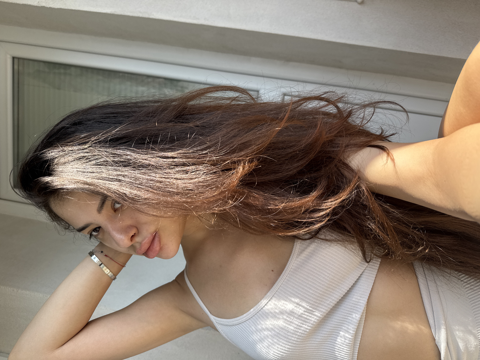 A young adult female is depicted in a medium shot indoors, positioned next to a window with strong natural sunlight casting highlights and shadows across the scene. She has long, dark brown hair with visible auburn and reddish tones, styled with volume and flowing over her left shoulder and arm. Her face is turned slightly towards the viewer, with a direct gaze and a slight pout on her full lips, which appear to have a light pink or peach tint. She has a small, gold-colored nose ring and distinct eyebrows. Sun glints are visible on her forehead and nose.
She wears a light-colored (off-white or beige) ribbed crop top that reveals a portion of her midriff, and matching ribbed bottoms, the waistband of which is visible in the lower left of the frame. Her left arm is raised and bent at the elbow, with her hand positioned behind her head or within her hair. On her left wrist, she wears a silver-colored bracelet featuring screw motifs, resembling a Cartier Love bracelet, and below it, a thin red string bracelet with a small dark bead. Her right arm is partially obscured by her hair and body.
The background consists of a plain, light-colored (white or off-white) wall on the left side and a white-framed window on the right. The windowpane reflects light and shows indistinct, blurred vertical elements that may be blinds or an distant outdoor view. There are no identifiable architectural features, landmarks, or unique elements within the image that allow for the specific geographic location to be determined as Plovdiv, Bulgaria, or any other city.