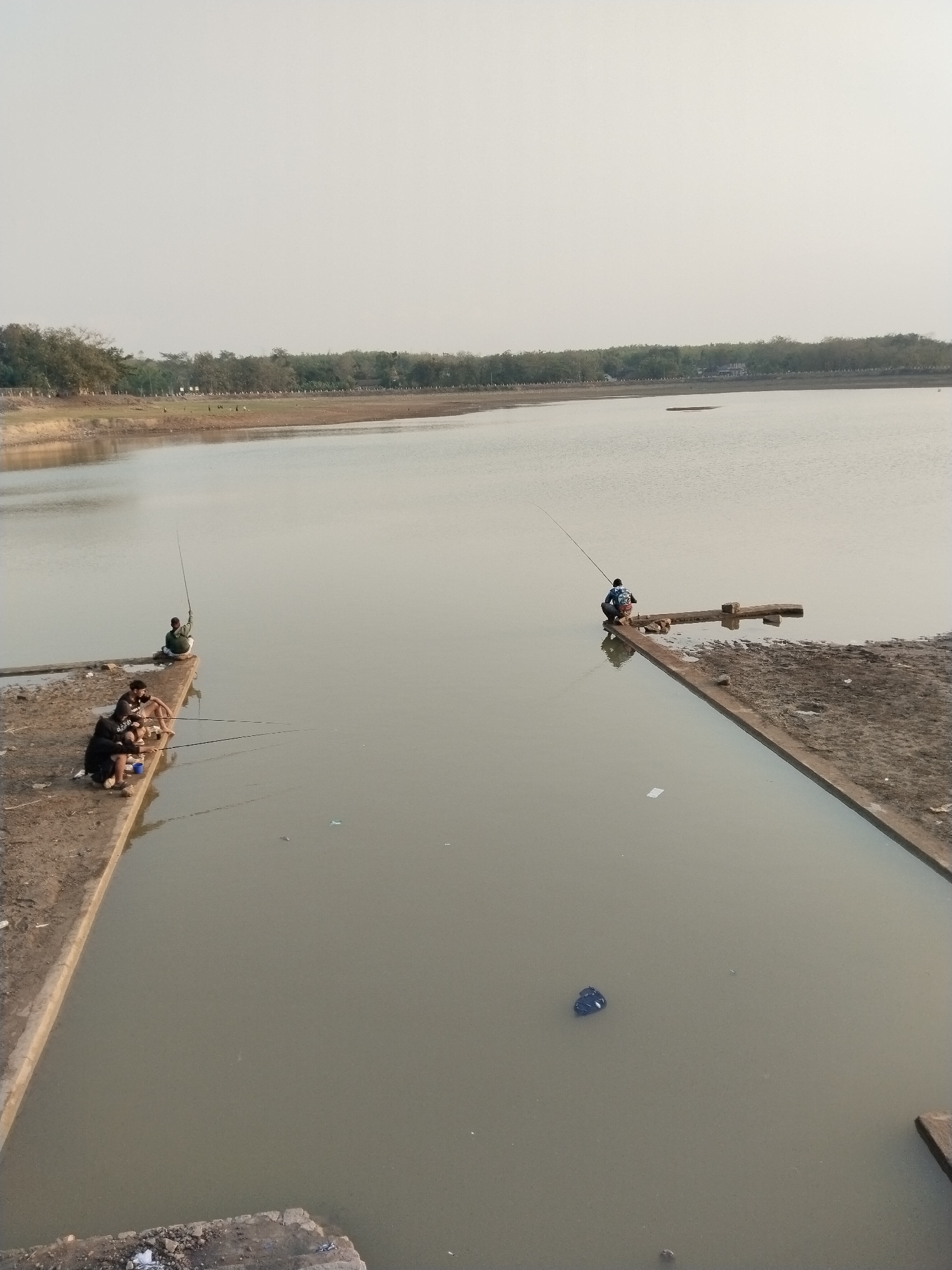 The image depicts a body of water with a muddy shore and a line of trees in the background. In the foreground, four individuals are fishing from a concrete structure extending into the water. Two of the fishermen are seated together, while the other two are positioned further apart. The water is calm and reflects the overcast sky. The overall scene suggests a quiet, possibly rural or park-like setting in Blimbing, Indonesia, where people are engaged in recreational fishing.