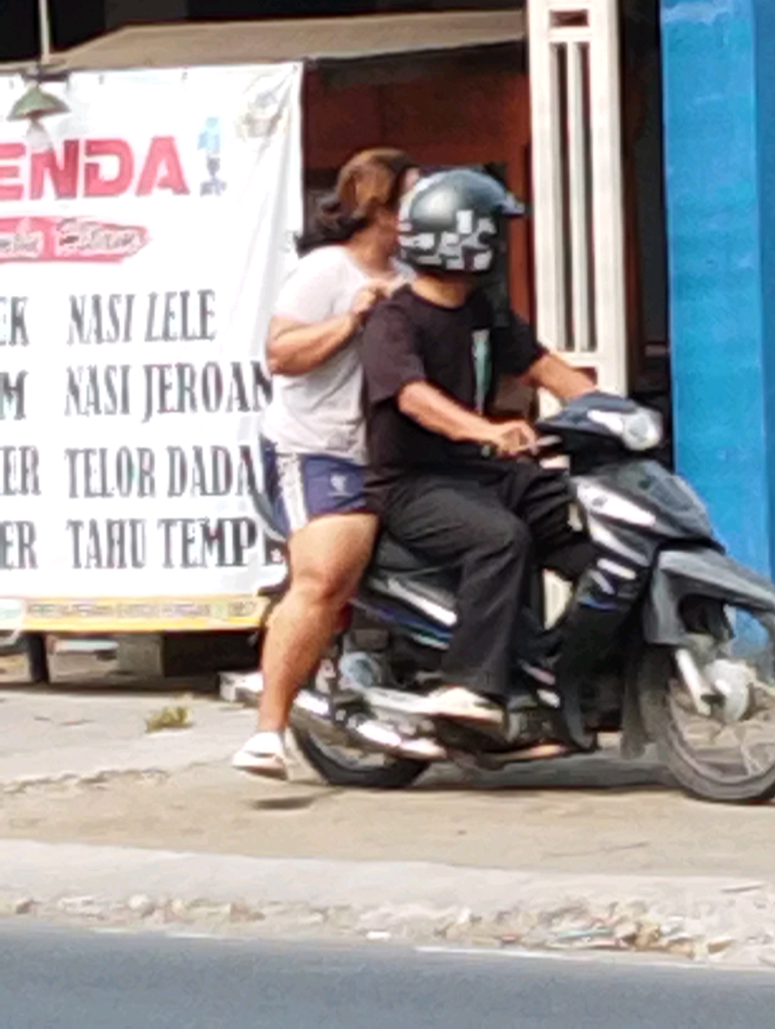 In Manaruwi, Indonesia, two individuals are pictured on a motorcycle. A person wearing a black t-shirt and black pants, with a patterned helmet, is operating the motorcycle. Behind them, another person in a light-colored t-shirt and blue shorts is seated. The motorcycle is dark-colored. Behind the people, a white banner is visible with red lettering that reads "ENDA" and other Indonesian text, possibly indicating food items. The environment appears to be an outdoor setting with a paved ground surface.