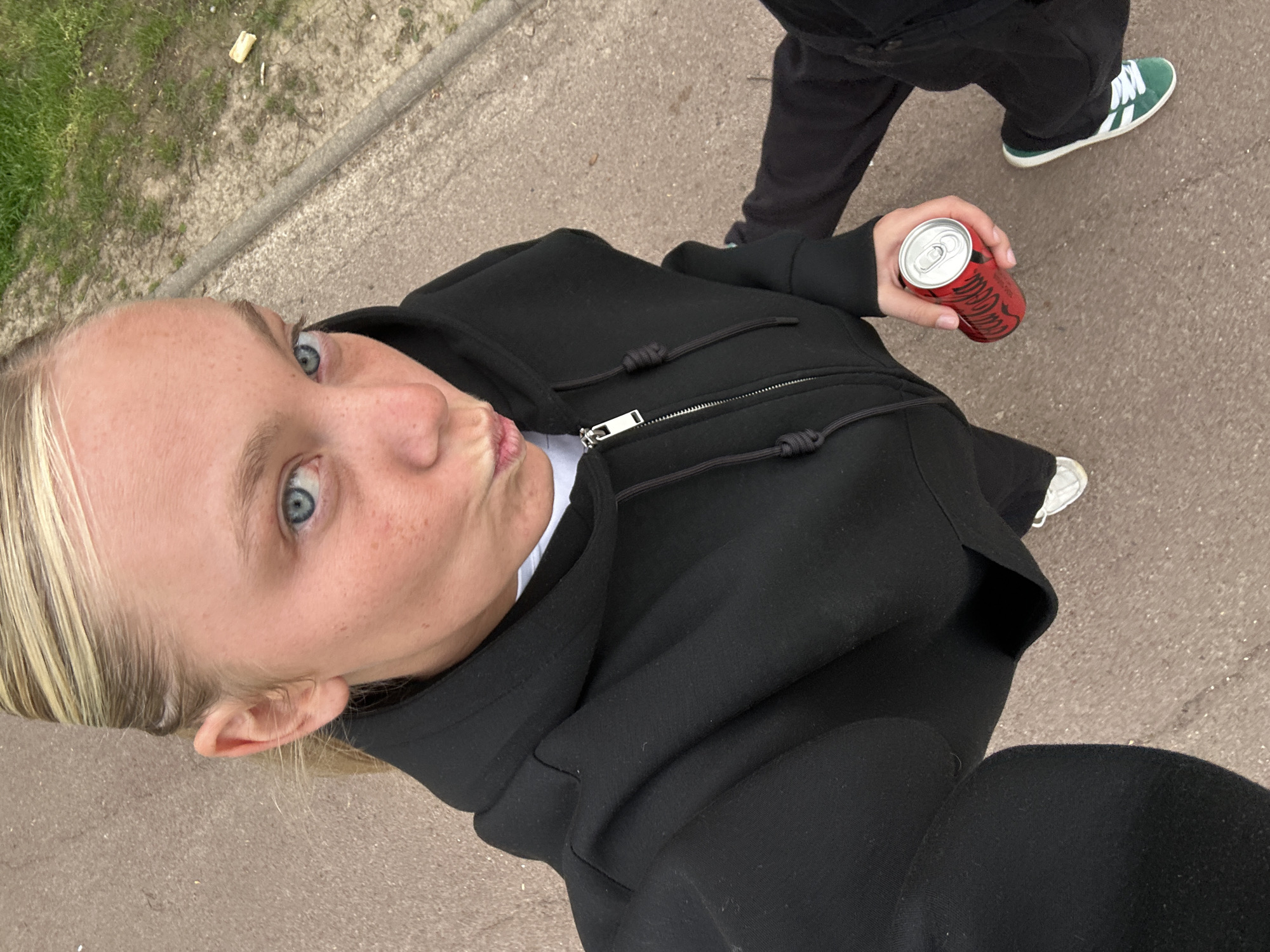 A woman takes a selfie while walking down a street with a person. The woman has blonde hair, fair skin, and blue eyes. She is wearing a black hoodie with a white t-shirt underneath. She holds a red can with a silver top in her right hand. In the background, a person walks with the woman. This person wears black pants and green and white Adidas sneakers. The sidewalk is made of gray concrete and has some green grass. The location is unknown.