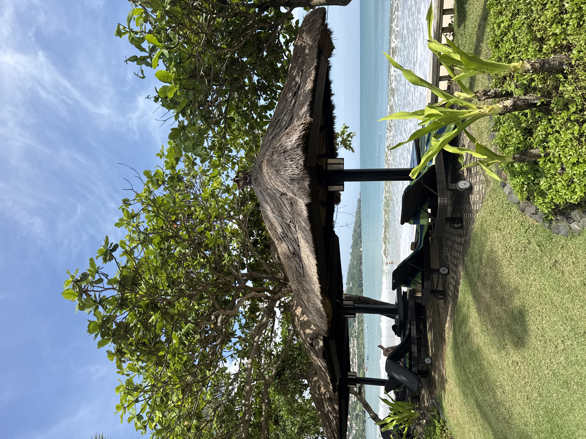 The image presents a view of a beach scene in Kuta Selatan, Indonesia. It appears to be taken from a slightly elevated position, likely from a veranda or similar structure. In the foreground, there is a thatched-roof structure, possibly a beachside hut or gazebo, supported by dark pillars. Beach lounge chairs are arranged beneath it. To the left of the gazebo is a tree with dense green foliage, partially obscuring the structure. To the right of the gazebo, green plants are visible along the edge. The background shows the ocean, with waves breaking along the shore, where a few people are visible. A stretch of sandy beach separates the ocean from the green vegetation along the coast. The sky is blue with scattered clouds, suggesting a clear day.