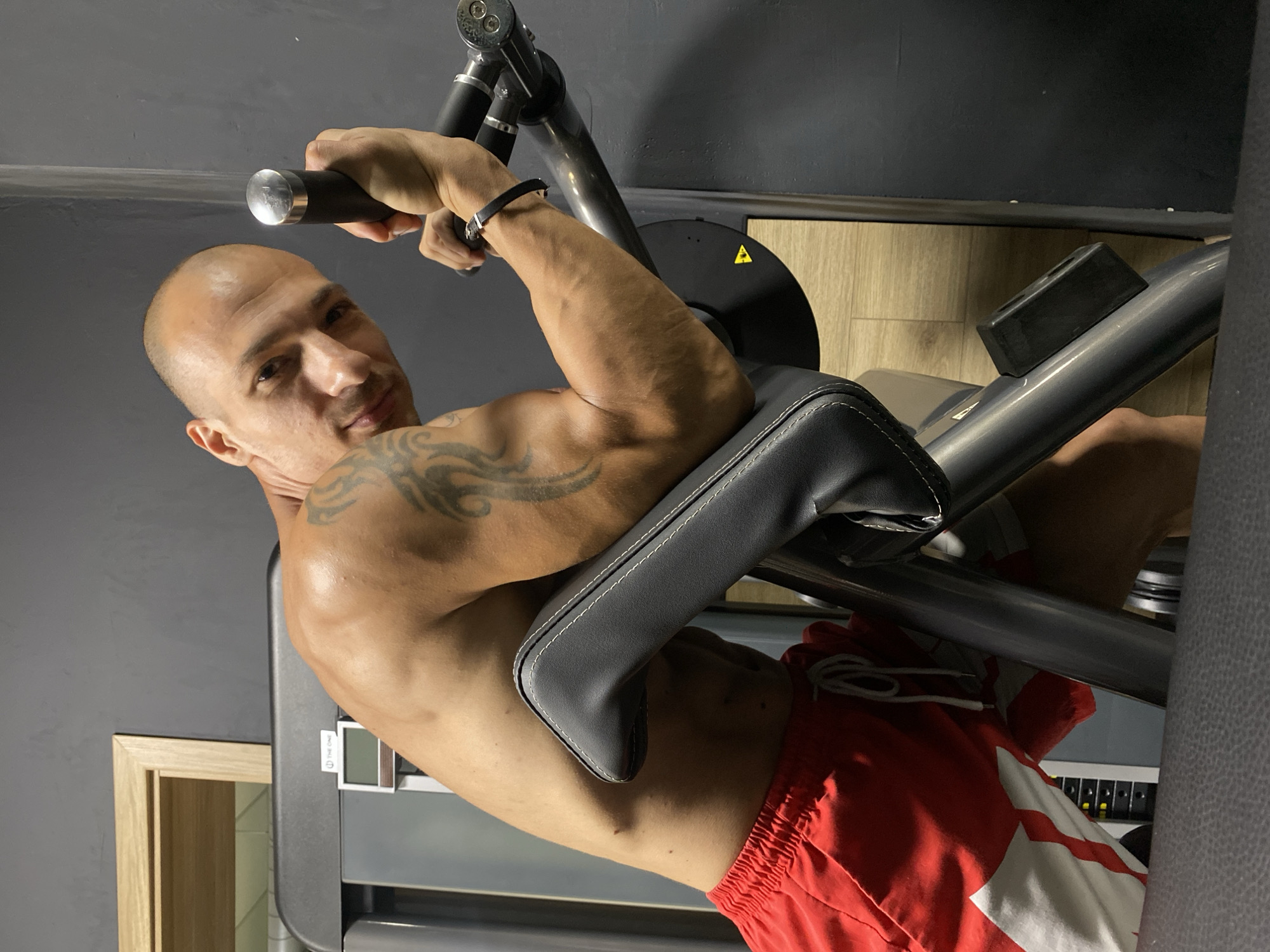 A bald man with a tattoo on his left arm is exercising in a gym. He is wearing red shorts with a white design. He is leaning on a gym machine with his left arm and gripping a handle with his right hand. The gym appears to have dark gray walls and wooden flooring. There is a wooden door frame in the background. The image was taken in Pleven, Bulgaria.
