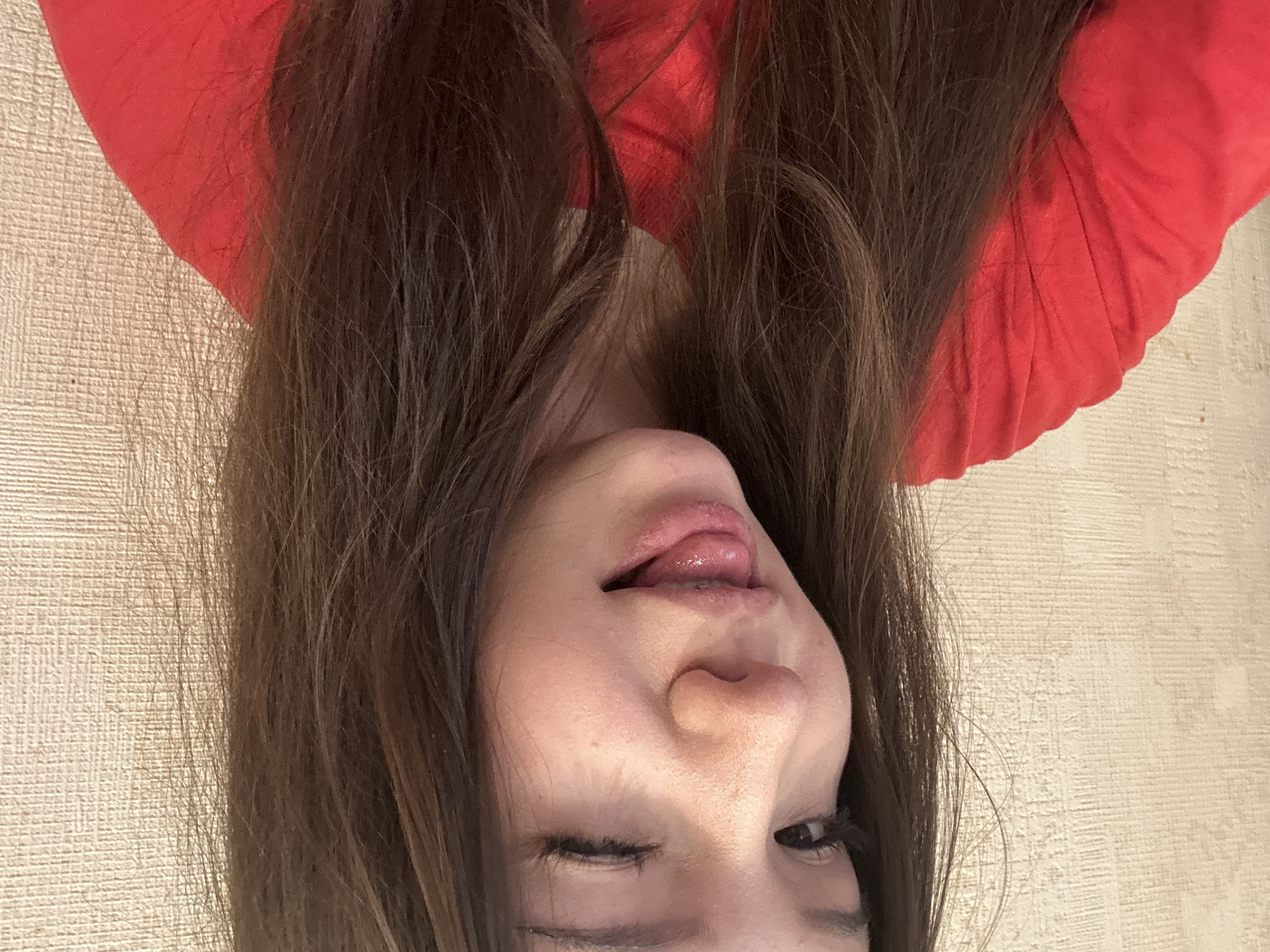 A young individual, appearing female, with long, dark brown hair, is captured in a close-up, chest-up shot. The subject is wearing a bright red, short-sleeved top. Their left eye (viewer's right) is winked shut, while the right eye (viewer's left) is open and directed towards the viewer. The mouth is slightly open, revealing the tongue extended slightly downwards and outwards, and visible braces on the lower teeth. The background is a plain, light beige or off-white wall exhibiting a subtle horizontal linear texture, suggesting wallpaper or textured plaster. The setting appears to be indoors. The image lacks discernible features to confirm a specific location within Naberezhnye Chelny, Russia.