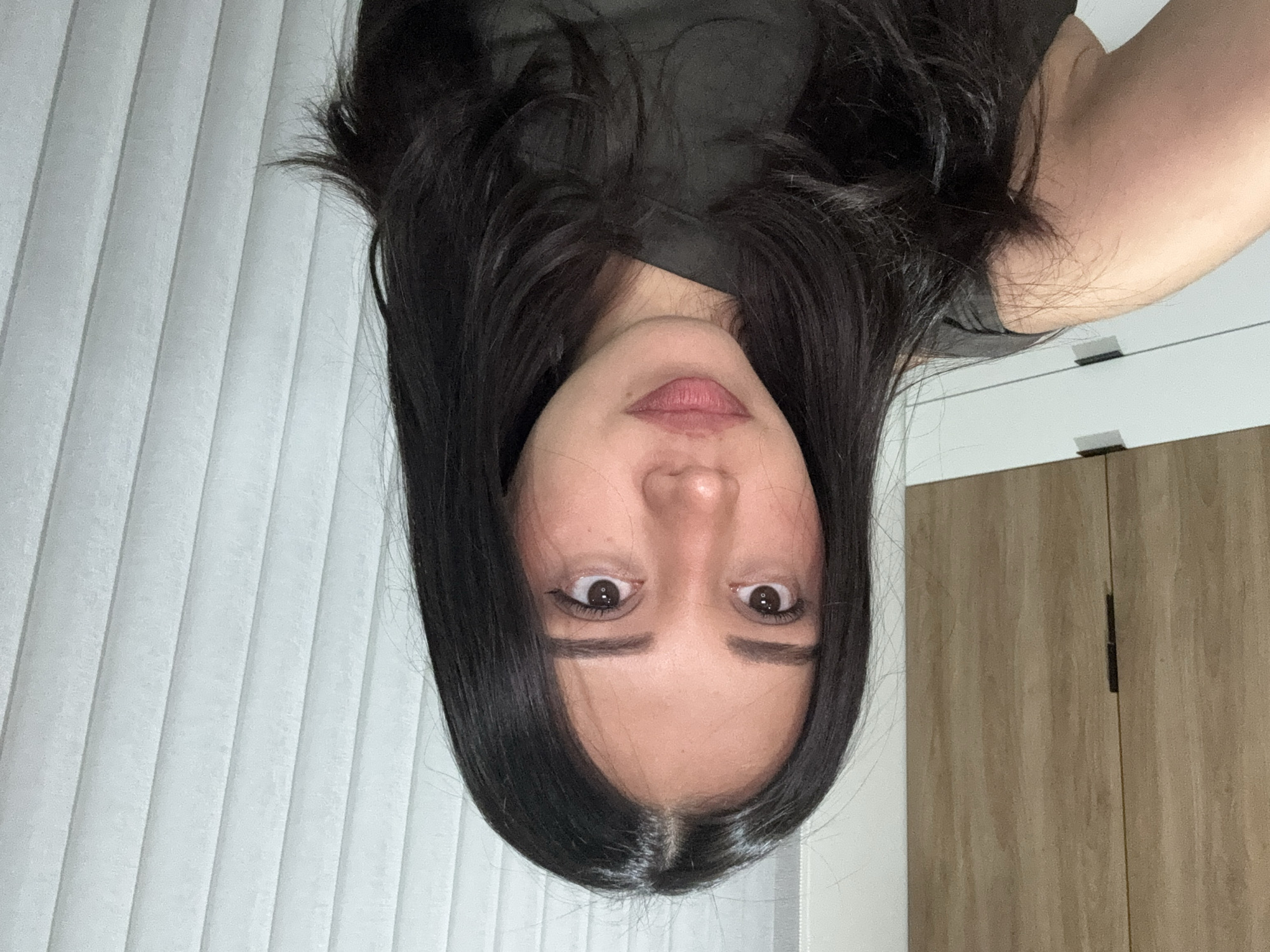 The image displays a close-up, indoor portrait of a single individual. The subject is a young adult female with a medium skin tone, dark brown eyes, and dark, straight, layered hair parted in the center, extending past her shoulders. She is wearing a dark, possibly olive green or charcoal-colored, short-sleeved top. Her gaze is directed slightly off-camera to the viewer's right, maintaining a neutral expression. The lower-left portion of her left arm is visible, consistent with the act of self-photography.
The background is divided into two sections. On the left, light brown wooden cabinetry with a vertical grain is visible, featuring a dark, rectangular handle on an upper cabinet door. Below this, white cabinetry with dark horizontal handles is present. On the right, white vertical blinds are prominently featured. The lighting is even and consistent with an interior setting.
The specific location within the city of Puebla, Mexico, cannot be determined from the visual information presented in this image.