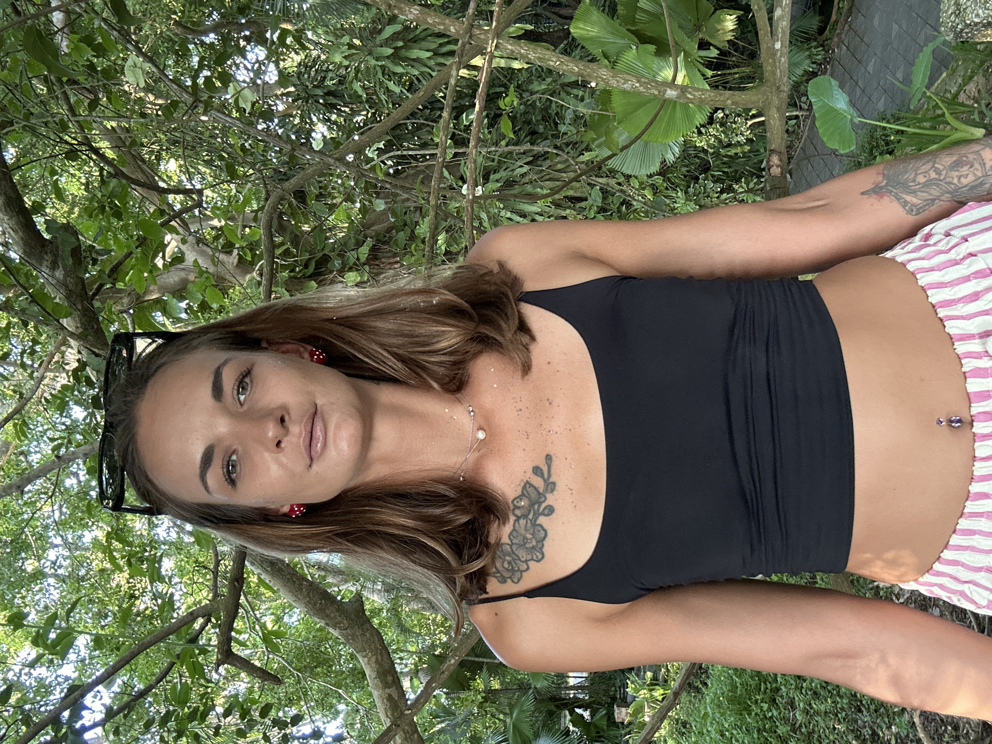 A woman with brown hair and green eyes is captured in a close-up, outdoor portrait. She is wearing a black tank top and striped shorts. A tattoo of flowers is visible on her chest, and a belly button piercing is also present. She has red and white mushroom-shaped earrings. Sunglasses are perched on her head. The background is lush with green foliage, suggesting a tropical setting, possibly in Phuket City Municipality, Thailand. The lighting indicates it is daytime, with dappled sunlight filtering through the leaves. The woman's expression is neutral.
