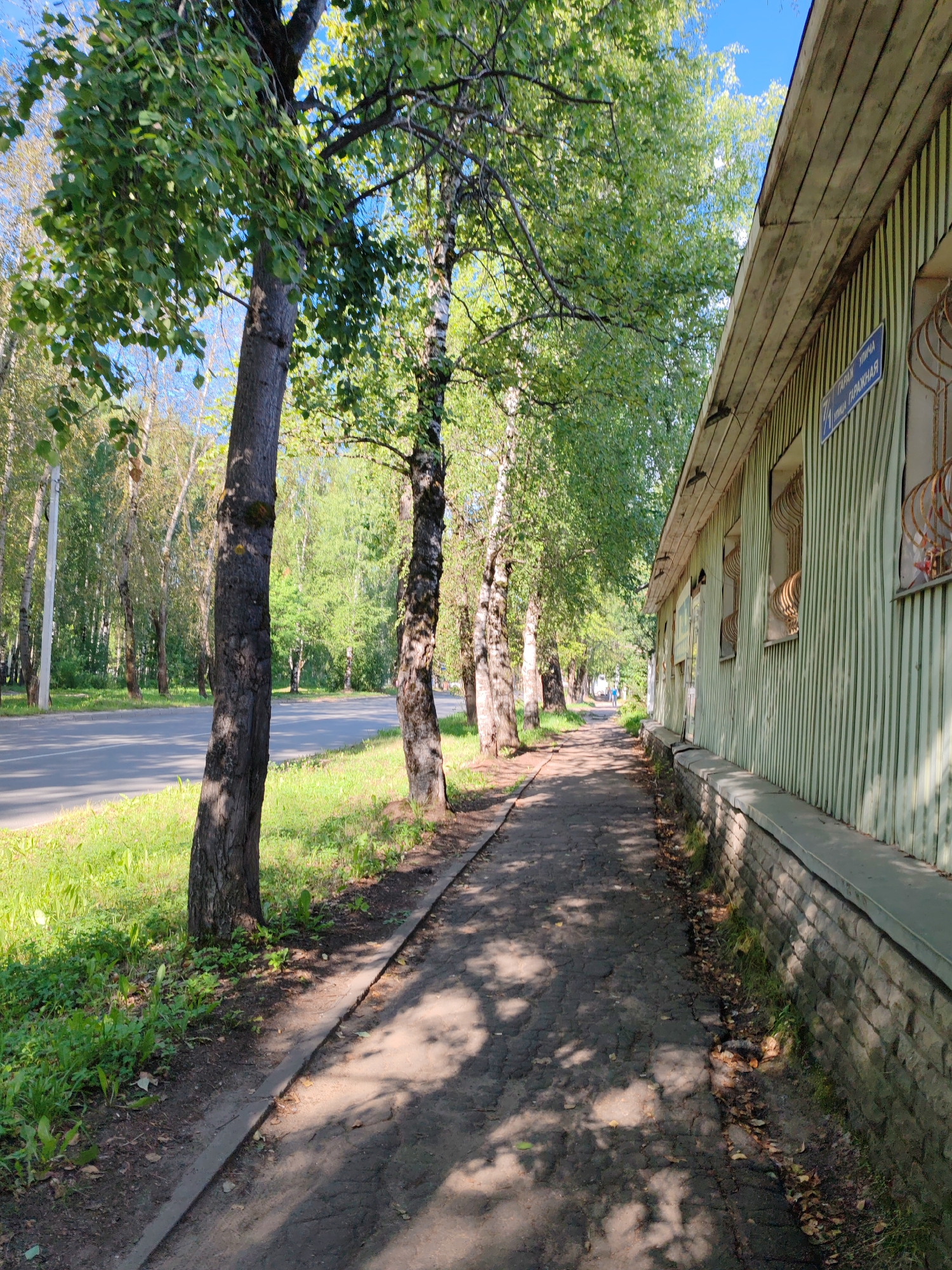 This is a full outdoor shot of a sidewalk along a street in Syktyvkar, Russia. On the right side of the image is a light green building with a wooden roof and a stone foundation. The building has several windows with decorative metal bars. A street sign that reads "ULITSA TARANOV GRANCHNAYA" is visible on the building's wall. The sidewalk is paved with asphalt, and is lined with trees on the left. The street is on the left side of the image, with grass and trees on the other side. Sunlight casts shadows across the sidewalk and street. There are no people visible in the image.