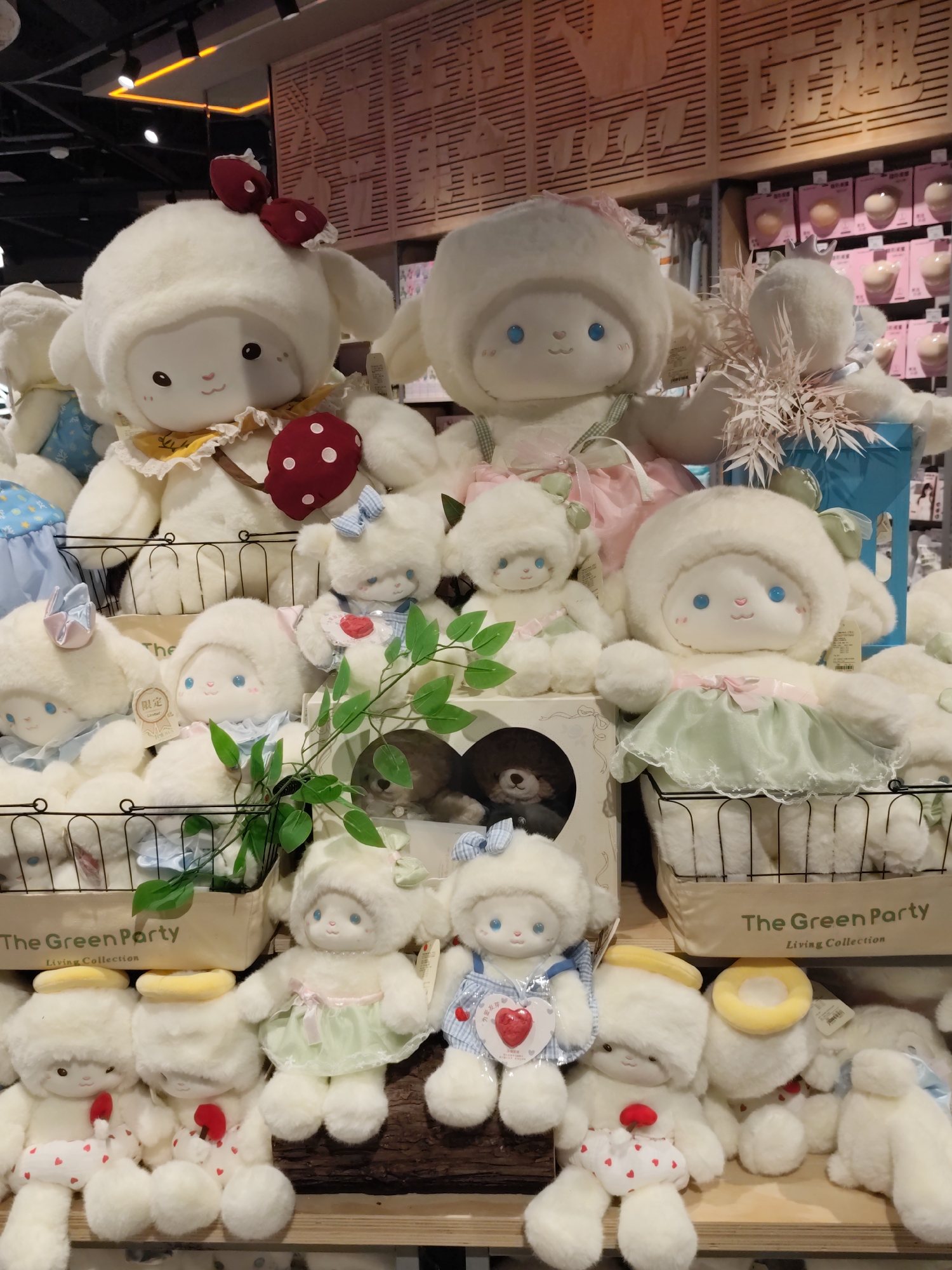 The image shows a display of plush toys, primarily white sheep or rabbit-like characters with large, round heads, blue eyes, and small pink noses. The toys are arranged on shelves in what appears to be a retail environment. Several of the toys are wearing clothing, including dresses, overalls, and hats. Some are adorned with bows, flowers, or halos. In the background, there are shelves stocked with various products, and the overall lighting suggests an indoor commercial space. Text on a tan box in the middle foreground reads "The Green Party Living Collection." While there are no people visible in the image, the scene is characteristic of a store display. This display is located in Daqing, China.