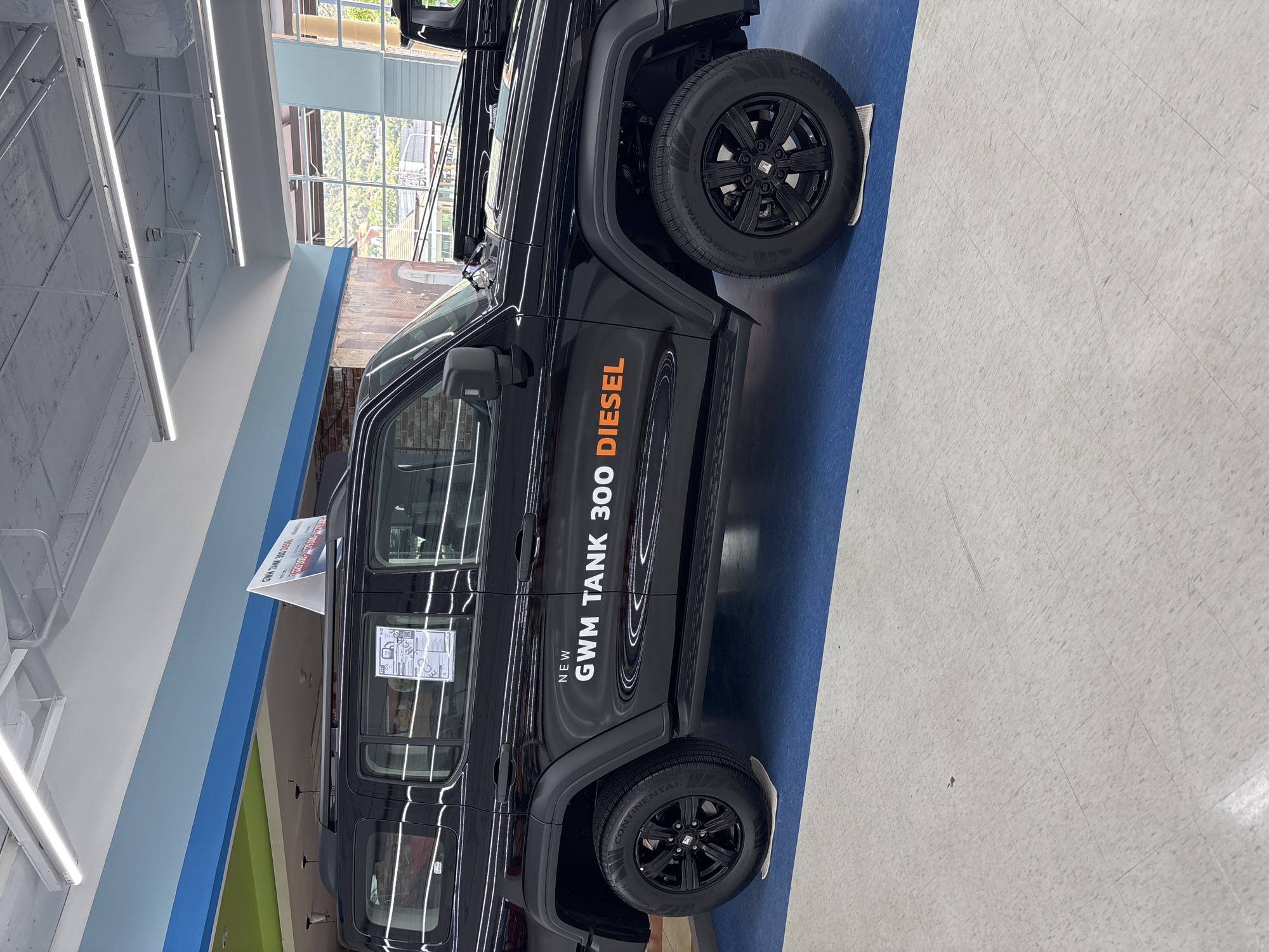 The image shows a black GWM Tank 300 Diesel car. The car is parked indoors on a blue and gray floor in an area with overhead fluorescent lighting. The side of the car is visible, showing the text "NEW GWM TANK 300 DIESEL". There is a white sign visible on the car's side window. The location, บ้านบางรักษ์, Thailand, cannot be identified from the visible elements in the image.