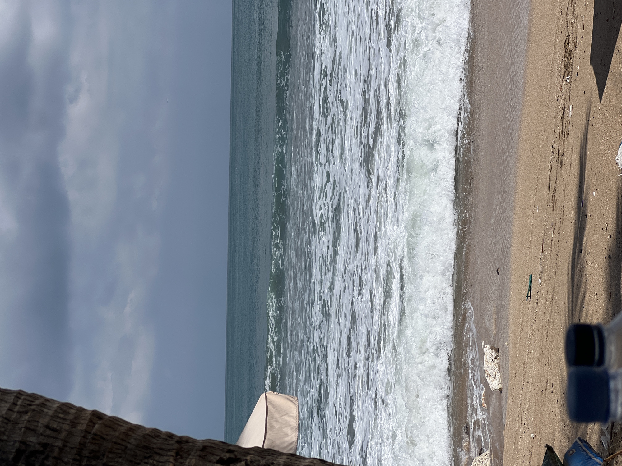 The image depicts a natural scene at a beach in Kuta, Indonesia. A large tree trunk is visible on the left side, partially obscuring a beach umbrella with a beige canopy. The ocean stretches across the frame, with waves breaking on the shore. The water is a deep teal color, and the sky above is overcast with gray clouds. The beach itself is sandy, with some scattered rocks and debris. In the foreground, two plastic bottles with blue and black caps are visible, partially out of focus. The overall impression is one of a somewhat cloudy day at a tropical beach.