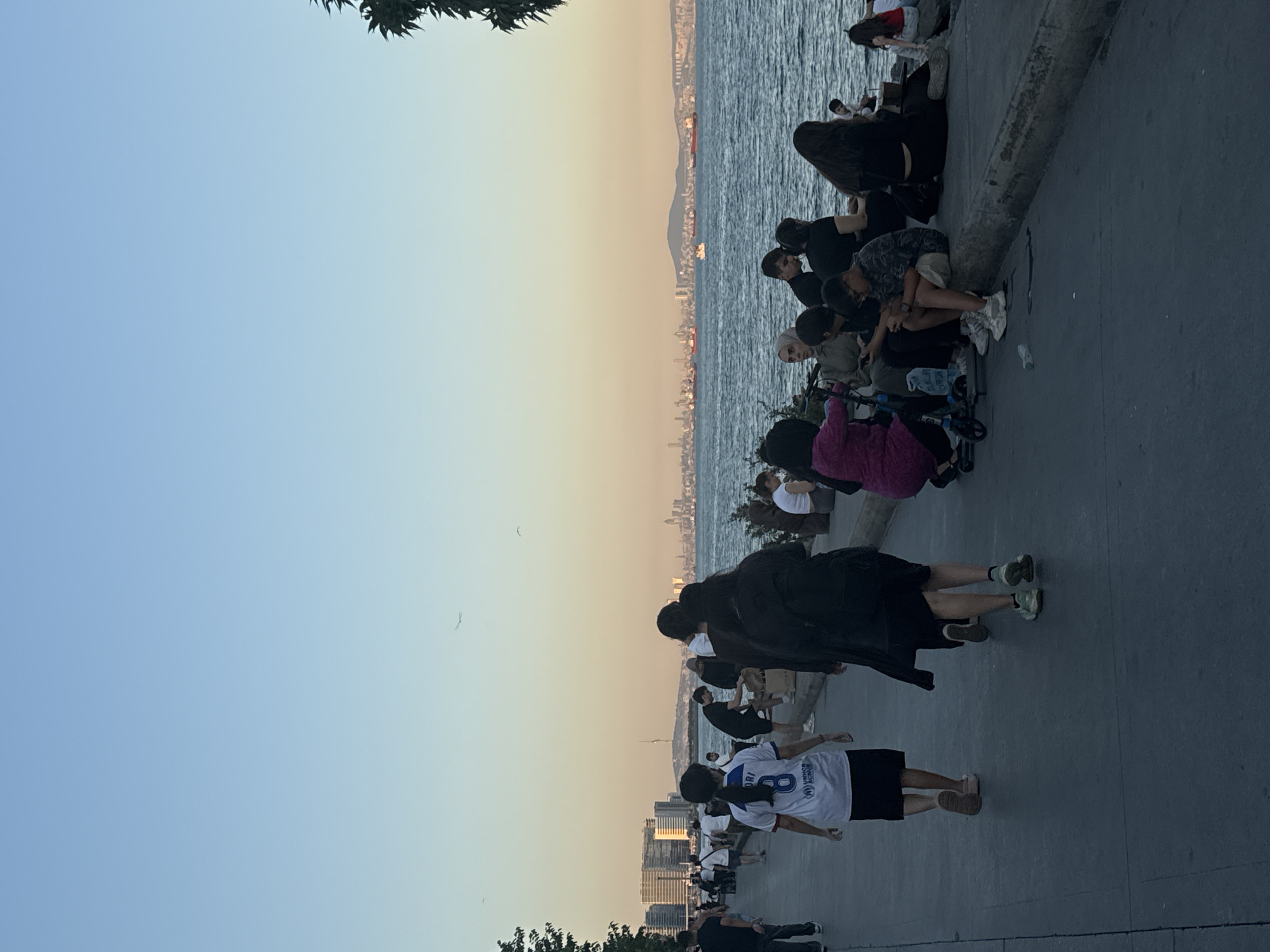 The image captures a waterfront scene in Bakırköy, Turkey, with a promenade bordering a body of water. The sky transitions from a clear blue at the top to a pale yellow near the horizon, indicating late afternoon or early evening. In the background, a distant city skyline is visible, punctuated by buildings and what appears to be a radio tower. Several cargo ships are anchored in the water.
On the promenade, a number of people are present. In the foreground, a young woman wearing a white and blue sports jersey with the number '8' and the UNHCR logo walks away from the camera, accompanied by another person in black attire. Further down the promenade, more individuals are walking, including a man in a black t-shirt and shorts.
A group of people are seated along the edge of the water, on a concrete barrier. Some are looking at their phones, while others are engaged in conversation. A child is visible with a scooter near the seated group. The overall atmosphere suggests a leisurely gathering by the sea.
