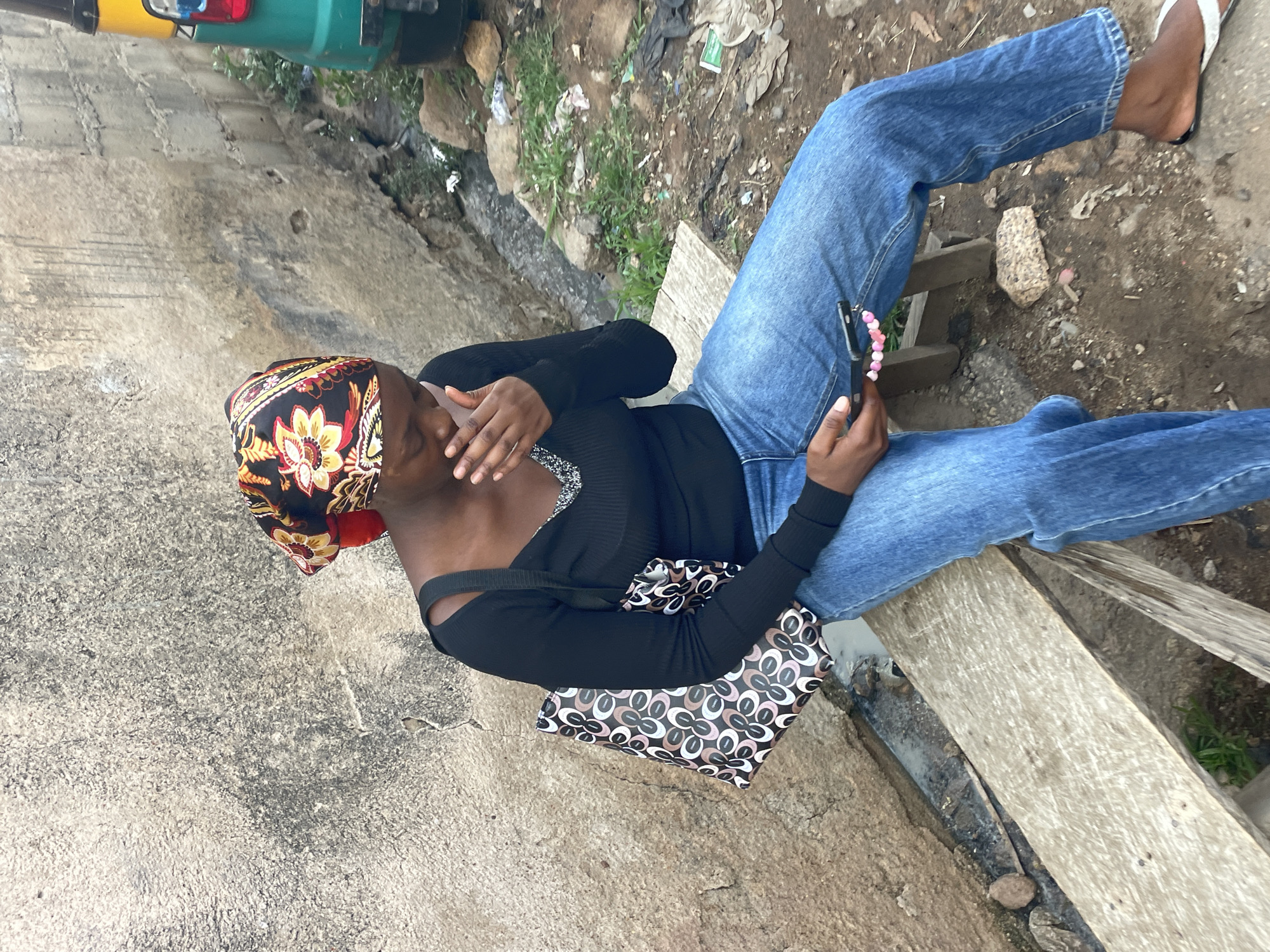A woman sits outdoors in Jos, Nigeria, on a makeshift wooden bench. She wears a headscarf with a floral pattern, a long-sleeved black top, and blue jeans. She holds a phone with a beaded charm in her right hand, and her left hand is raised toward her face. A patterned purse rests beside her. The ground is concrete and partially covered with debris and sparse vegetation. A green and yellow vehicle is partially visible in the upper left corner.