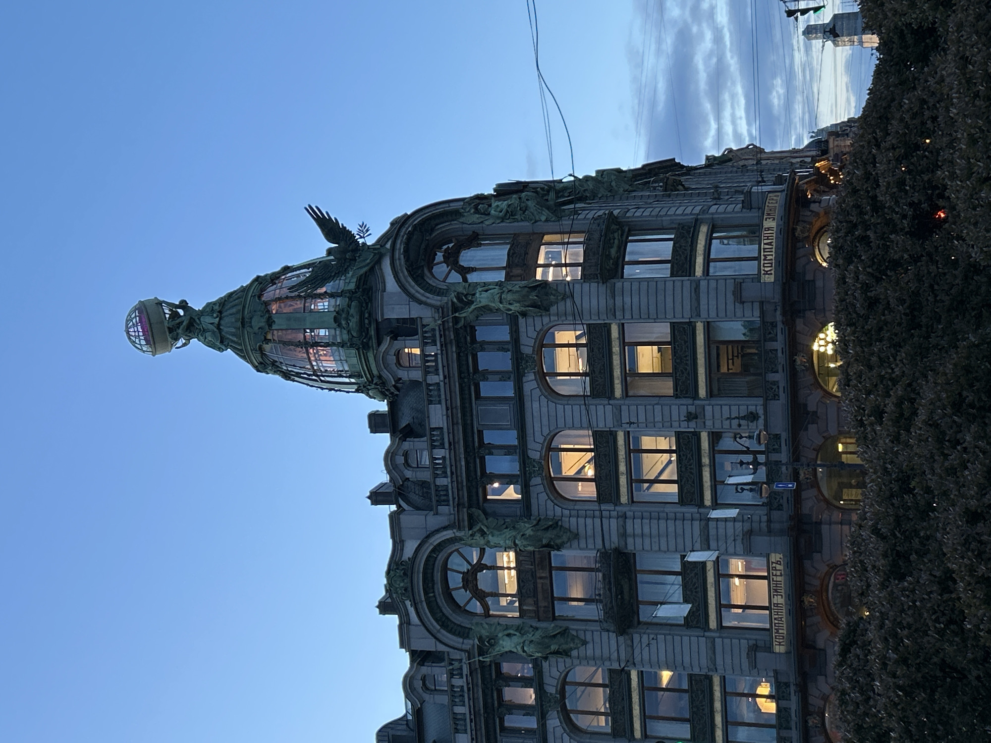 This media file presents a photograph of an ornate building against a twilight sky, rotated 90 degrees clockwise.
The primary subject is a multi-story building, likely an architectural landmark. Its facade is a light grey stone, richly adorned with intricate Art Nouveau-style decorations, including statues and relief work. A prominent feature is a multi-tiered, green-patinated glass dome or spire located on the leftmost side of the image (which would be the top when the image is viewed upright). This spire is capped with a sphere resembling a globe, and a sculpture of a bird with outstretched wings is visible near its base.
The building features numerous windows of varying shapes, including arched and rectangular designs. Many of these windows are illuminated from within, casting a warm, golden glow that contrasts with the exterior's cooler tones.
The sky, occupying the upper left portion of the image, is a clear, light blue, suggesting either dawn or dusk. Some faint, wispy clouds are visible towards the right edge of the sky. Several thin wires traverse the sky above the building. On the rightmost side of the image, a dense, dark green bush or tree obscures the lower part of the building.
No specific activity or event is taking place; the image is a static architectural study.
A notable detail is the visible text on the building's facade: "КОМПАНИЯ ЗИНГЕРЪ" (KOMPANIYA ZINGER), which translates to "SINGER COMPANY". This, combined with the distinctive dome and globe, strongly indicates the building is the Singer House (Dom Knigi) in Saint Petersburg, Russia. The time of day appears to be twilight, with the internal lights creating a welcoming ambiance against the dimming natural light.