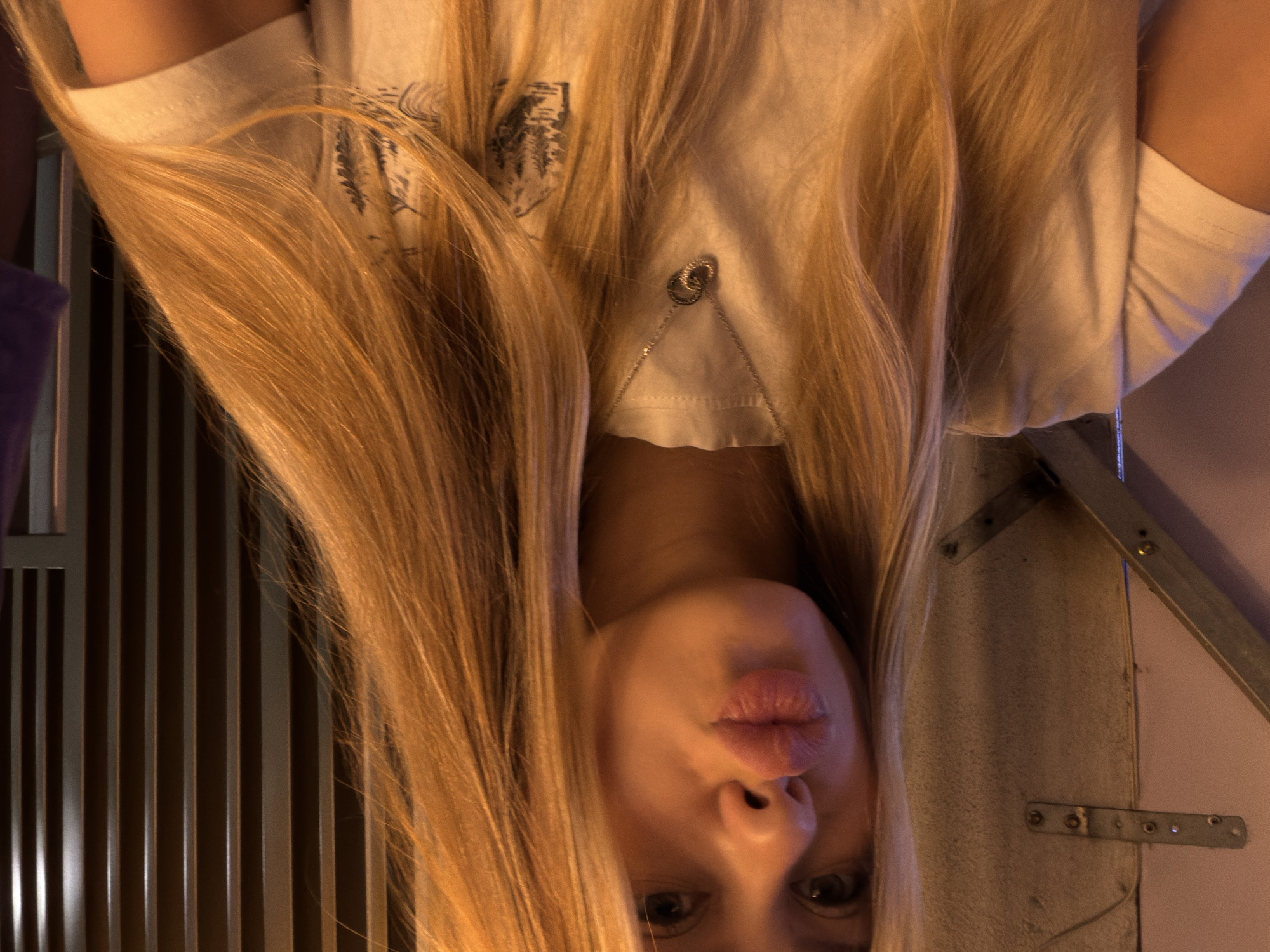 This media file is a close-up, upside-down portrait of a young woman with long, blonde hair. Her face is the central focus, with her eyes looking directly at the camera and her lips pursed in a kissing gesture. She is wearing a light-colored t-shirt with a subtle graphic print. A delicate silver necklace with interlocking rings is visible on her chest, resting on the fabric of her shirt.
The background is dimly lit and appears to be an indoor setting. To the left of the woman, there is a vertical slatted surface, possibly a room divider or a piece of furniture. To the right, a textured, light-colored surface is visible, with metal brackets and screws attached, suggesting it might be a wall or a constructed surface. The overall lighting is warm and somewhat diffused, with some harsh highlights, suggesting artificial lighting. The image has a slightly grainy quality, which could be due to low light conditions or the camera used. No specific time of day or weather conditions are discernible. The emotional tone of the image is playful and inviting, due to the woman's direct gaze and kissing pose.