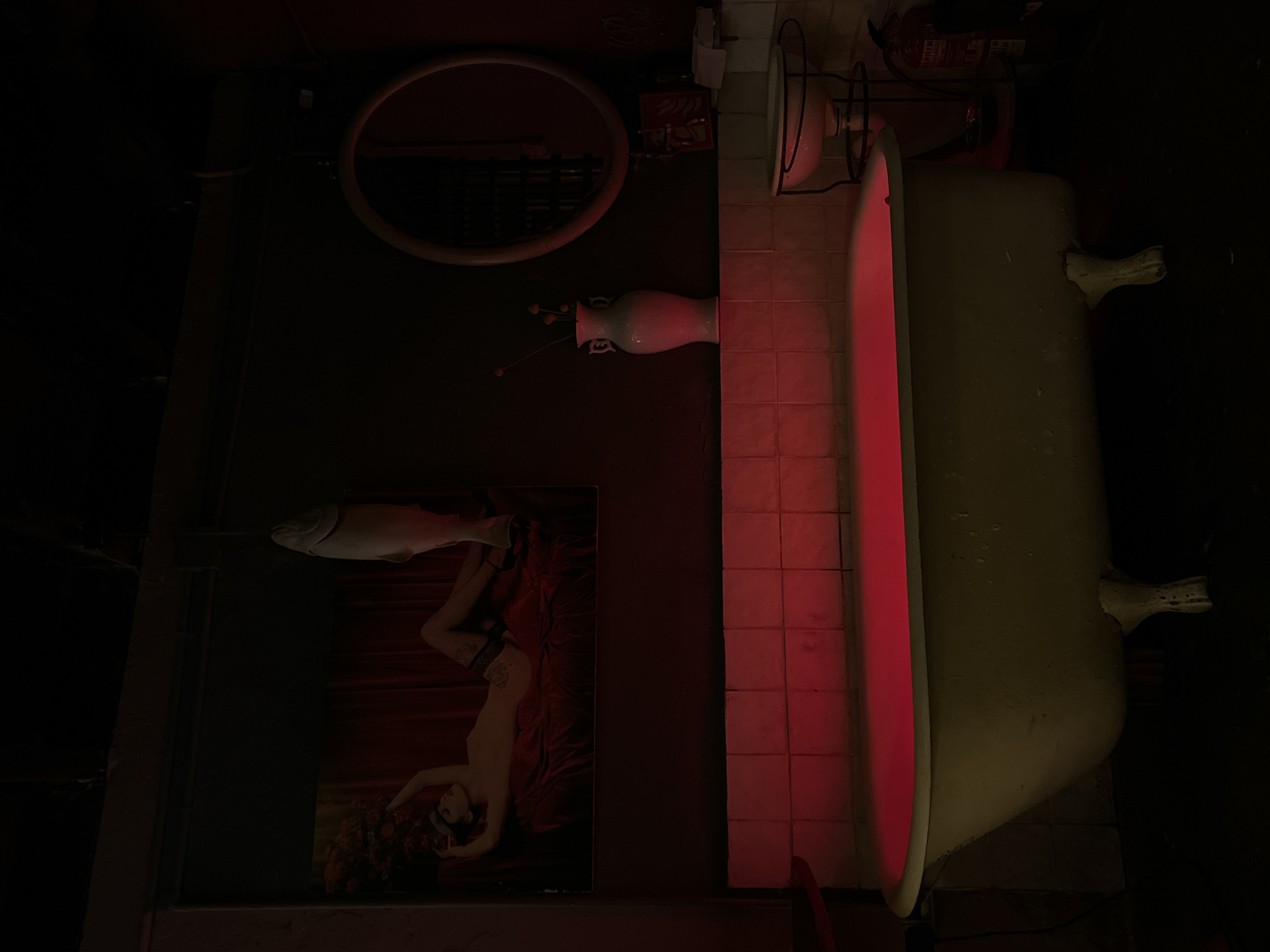 The image, if oriented correctly (rotated 90 degrees counter-clockwise from the provided view), depicts a dimly lit interior space with a striking red glow. The setting appears to be a uniquely decorated room, possibly a themed bathroom or an artistic installation, in what could be a commercial establishment like a bar or restaurant in Barcelona, Spain.
On the right side of the frame, a white, possibly claw-foot bathtub is prominently featured, emanating a strong red light from its interior. Behind the bathtub and extending to the left, there's a wall covered with light-colored square tiles. Mounted on this tiled wall, between the bathtub and other elements, is a red fire extinguisher.
Further to the left, a large, circular mirror with a thick, light-colored frame hangs on a darker wall. Below the mirror, a tall, slender white ceramic vase holds a few delicate, dried-looking stems or decorative sticks. Beneath the vase and mirror, a large framed artwork is displayed. This artwork depicts a reclining nude or semi-nude woman positioned on rich red drapery, with floral elements around her. An elongated, abstract white sculpture, resembling a fish, rests atop this framed picture.
No specific activity is taking place; the scene is static, showcasing the room's decor. The dominant red lighting creates a moody, artistic, and somewhat mysterious atmosphere, suggesting it's nighttime or an indoor space designed with specific ambiance in mind. Text is visible on the red fire extinguisher, but it is too dark and at an awkward angle to be clearly legible.