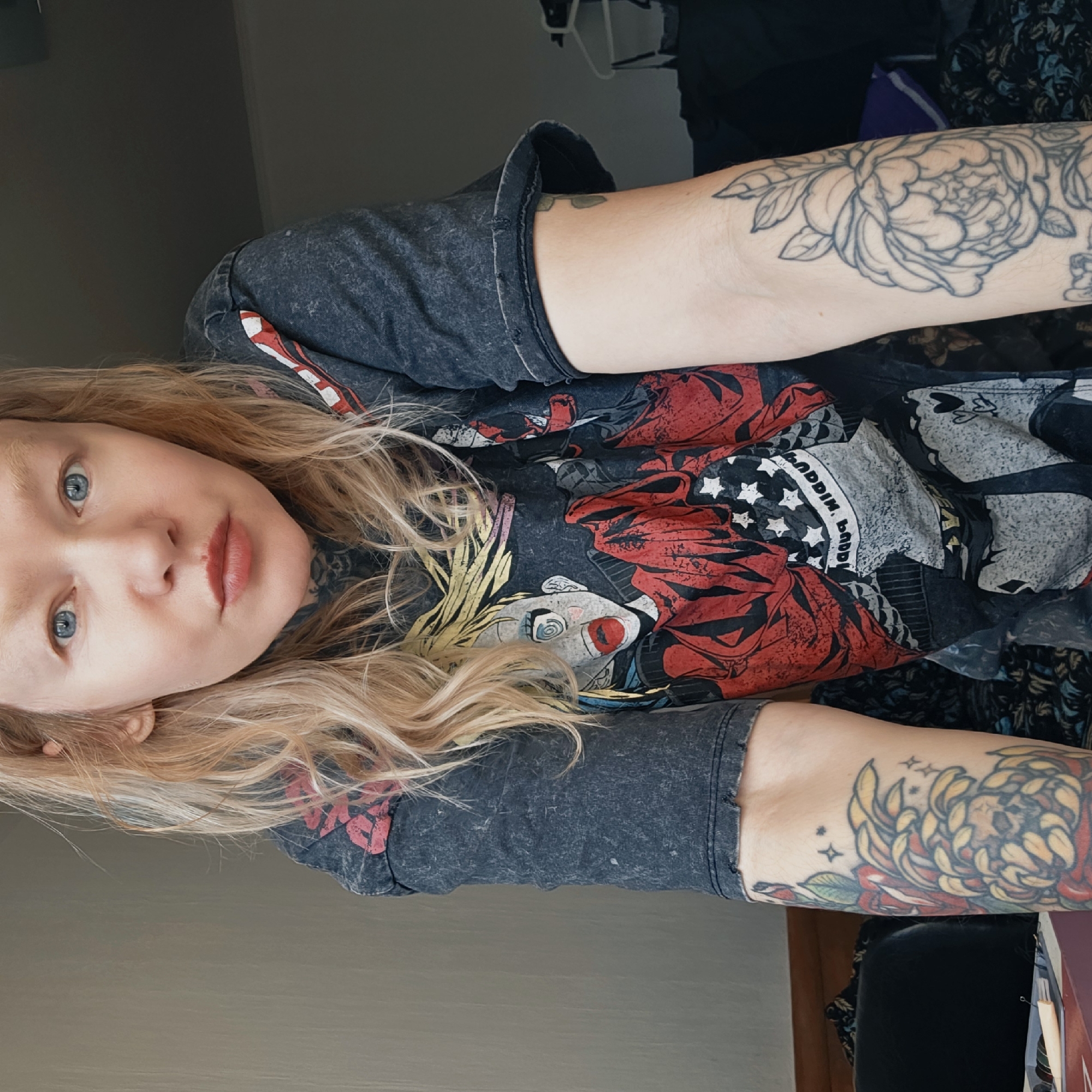 A fair-skinned person with light blonde, shoulder-length wavy hair and blue eyes is looking at the camera. They have tattoos on both arms, including a large floral tattoo on their right arm. They are wearing a dark-colored, printed t-shirt featuring a clown and text in a white font that says "ALADDIN, BABY". The background is muted and unclear, with hints of a wall and clothing. The location cannot be determined from the image.