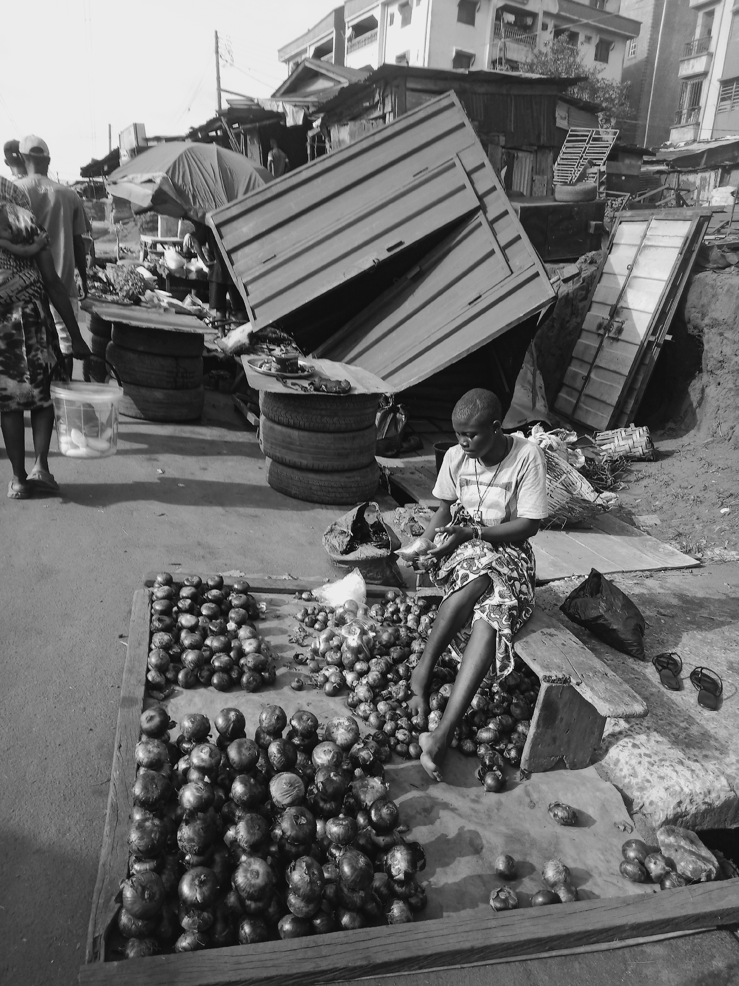 In a grayscale image taken in the city of Onitsha, Nigeria, a woman is seated on a makeshift wooden stool beside a large collection of onions spread out on a tarp within a wooden frame. She wears a patterned dress, a light-colored t-shirt, and a necklace. Her hands are occupied with small metallic objects. Behind her, a leaning metal structure dominates the background, with stacked tires serving as tables nearby. Two individuals are partially visible to the left, engaged in conversation. The surrounding environment is a cluttered street scene, with buildings of varying construction styles visible in the distance.