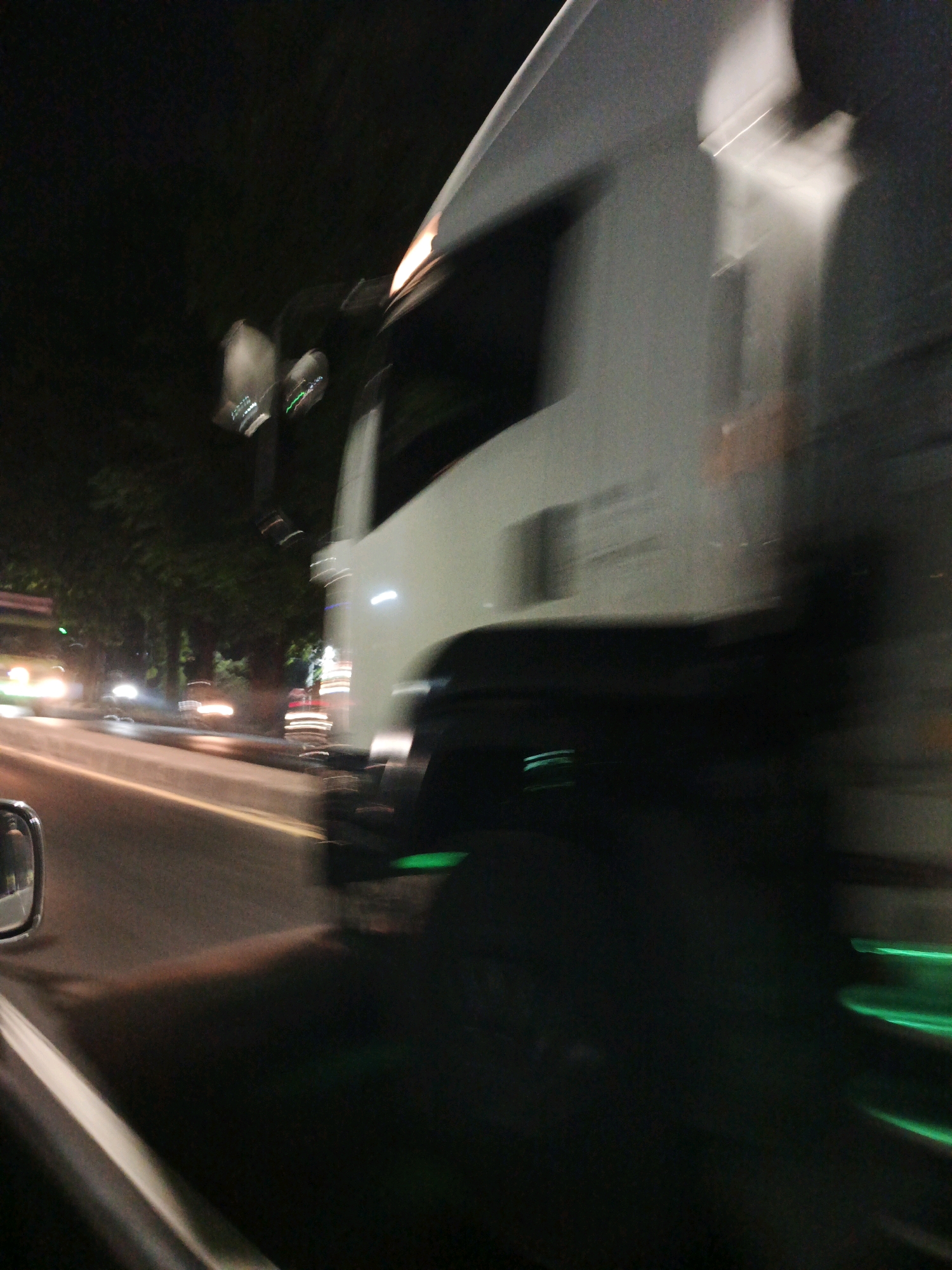 A blurred, nighttime street scene in Karangrejo, Indonesia, captured from the perspective of a moving vehicle. A large, white truck is prominently featured in the foreground, its details obscured by motion blur. The truck's headlights and side markers are visible as streaks of light. To the left, another vehicle with illuminated headlights and taillights is seen on the road, also blurred. The road surface is visible with white lane markings. Trees and foliage line the side of the road, appearing as dark masses against the night sky. Streetlights and other ambient light sources contribute to the illuminated environment. The overall impression is one of rapid movement through a city at night.