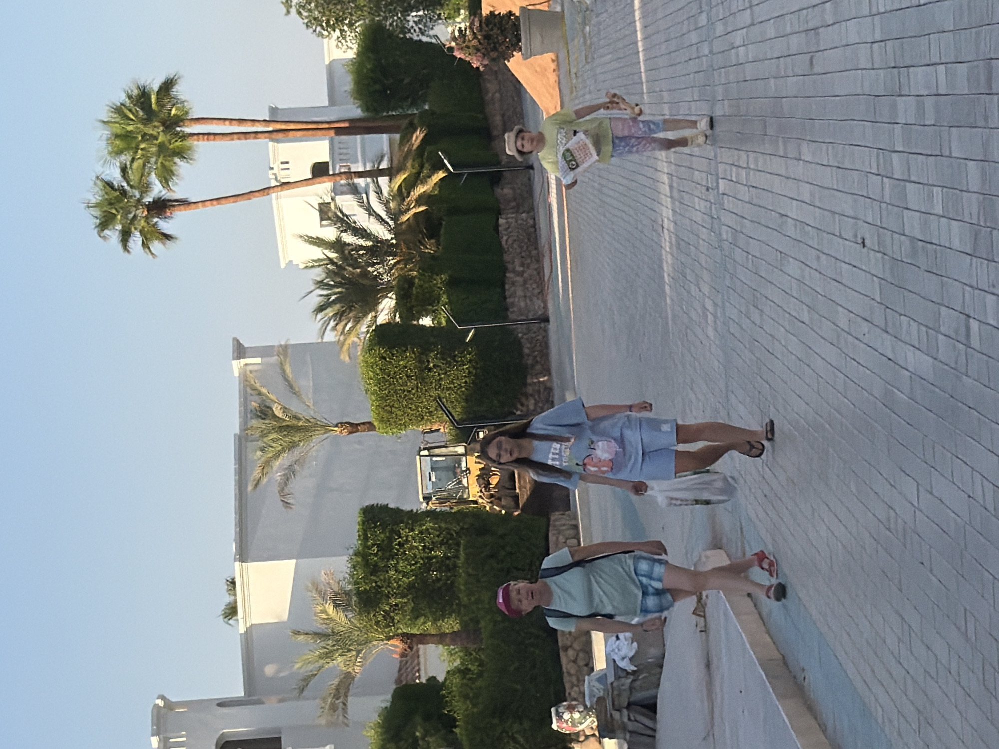 In an outdoor paved walkway in Sharm El Sheikh, Egypt, three individuals are walking. The first individual, on the left, is a woman wearing a pink hat, a light green t-shirt, and plaid shorts. She carries a dark backpack and is looking forward with her mouth slightly open. Beside her, to the right, is a young woman in a light blue t-shirt and light blue shorts, carrying a white plastic bag in her right hand and looking ahead. Further to the right and slightly behind is a child wearing a hat, a light yellow t-shirt, and multi-colored leggings. The child is holding a rolled-up object in their right hand and what appears to be a toy animal in their left hand, looking forward. In the background, there are buildings with white walls and distinctive architectural elements, some surrounded by lush green bushes and palm trees. A yellow construction vehicle is visible behind the bushes. The sky is clear and blue. The walkway is paved with grey brick-like stones.