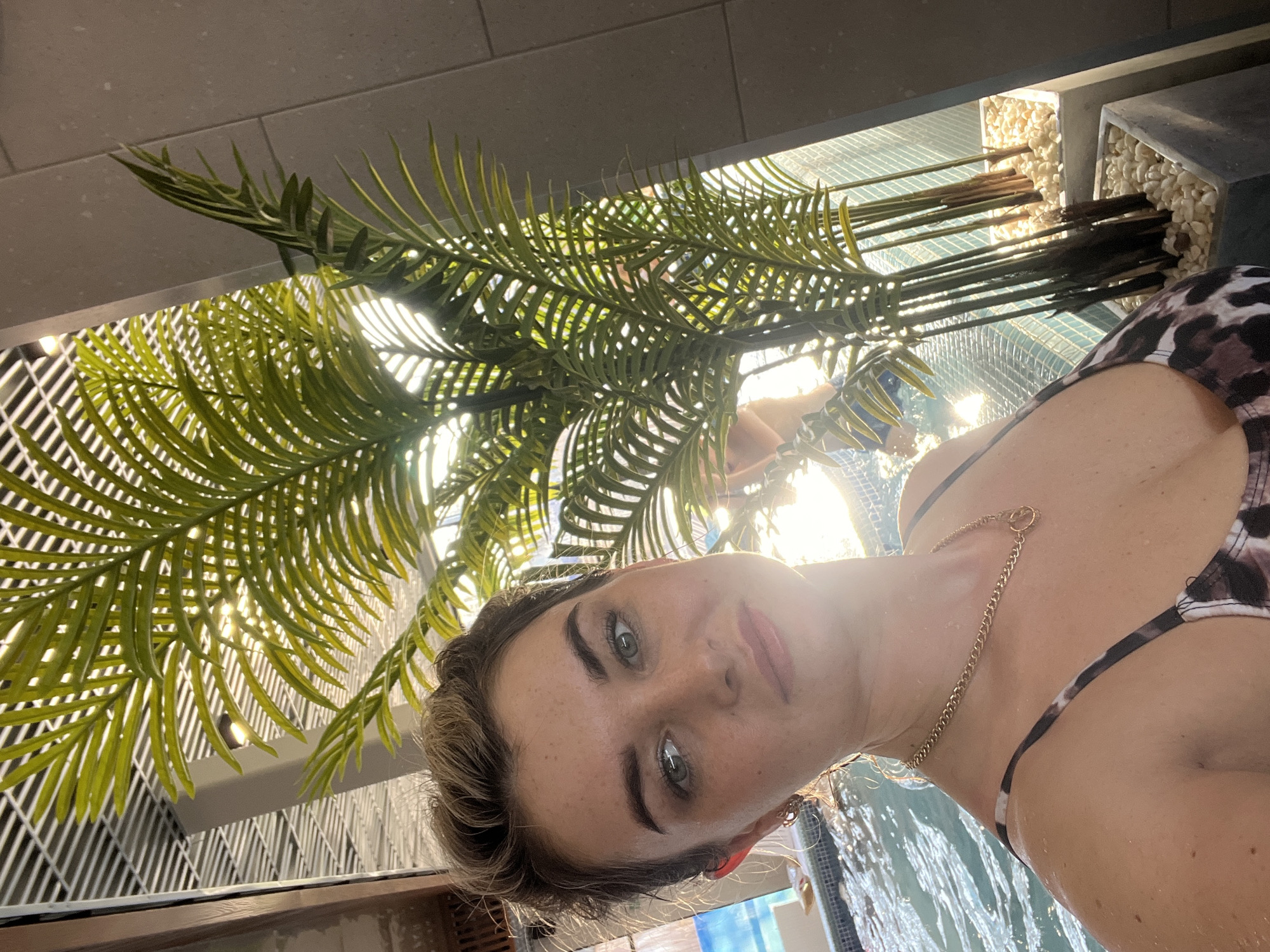 A woman is in a pool with green plants overhead. The pool has a tile bottom and a tile wall. The plants are in white pots filled with white pebbles. A chain necklace is visible on the woman. The location is not ascertainable as Votkinsk, Russia from the given information.