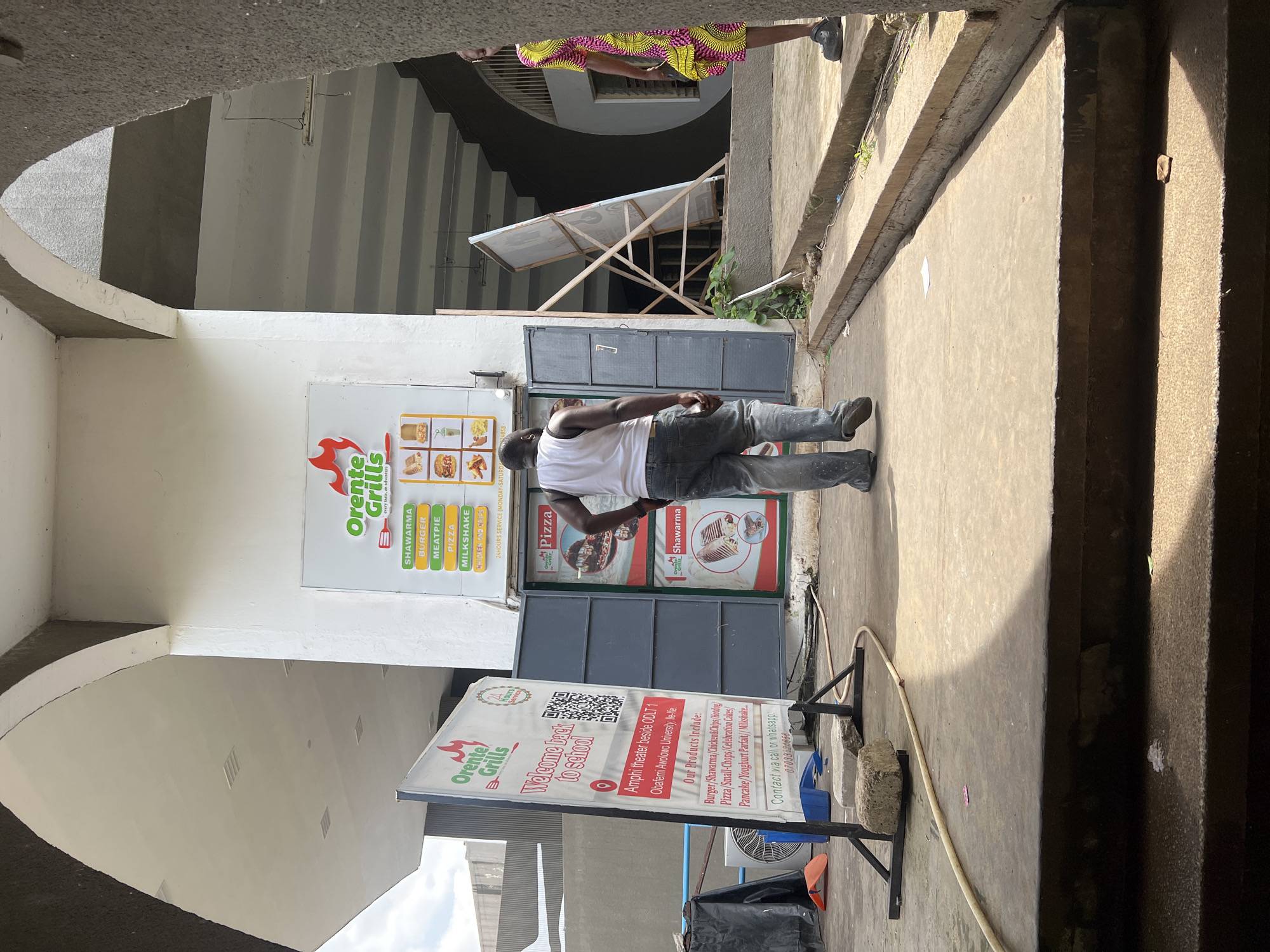 In the city of Ife, Nigeria, a man wearing a white tank top and gray pants stands with his back to the camera, facing a food stall. To his left, a white sign displays "Orente Grills" with the tagline "every taste, in signature." Below this are menu items like "SHAWARMA," "BURGER," "MEATPIE," "PIZZA," and "MILKSHAKE." Another sign on the left, also for "Orente Grills," features the text "Welcome back to school" and a QR code, followed by the location "Amphi theater beside ODLT 1, Obafemi Awolowo University, Ile-Ife" and a list of products including burgers, shawarma, pizza, and milkshakes. The stall itself has a gray metal door. To the right of the stall, a woman in a colorful, patterned dress is partially visible, facing away from the camera. The scene appears to be outdoors, under a covered walkway with concrete structures and stairs. The ground is paved concrete, with some debris and a coiled hose visible.