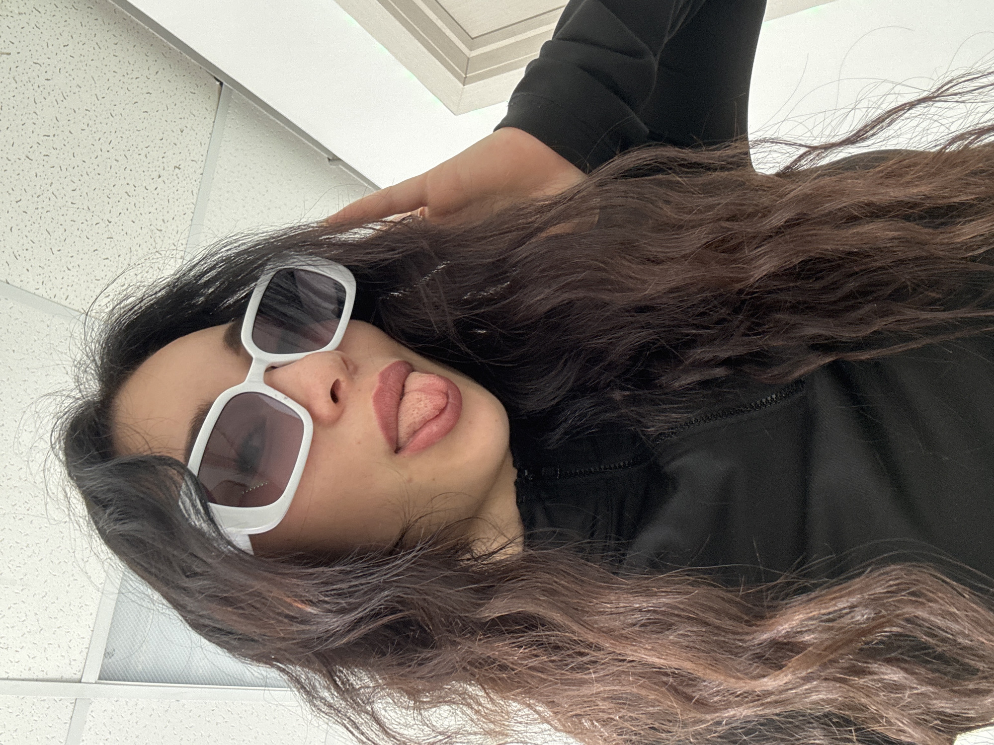 The image shows a woman with long, wavy brown hair, wearing white-framed sunglasses and a black jacket. Her tongue is slightly protruding. The background appears to be an indoor ceiling with square tiles. The location is not discernible from the image. There are no interactions or other subjects visible.