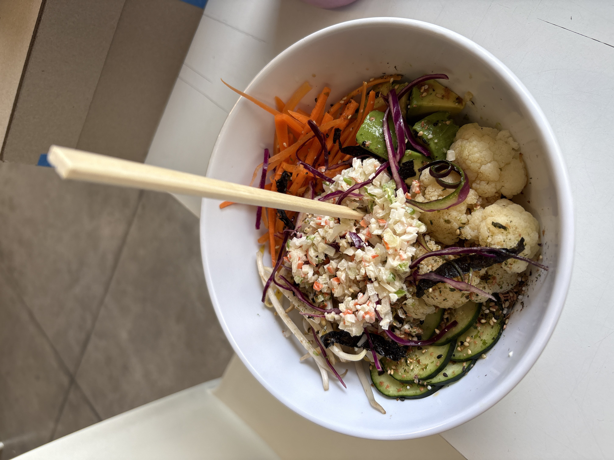 A high-angle, indoor shot displays a white bowl filled with a vibrant salad or grain bowl, held with chopsticks, on a white surface. Visible ingredients include shredded carrots, avocado chunks, cauliflower florets, sliced cucumbers, shredded red cabbage, bean sprouts, and a creamy salad mixture. The chopsticks are light brown and appear to be wood. The bowl is centrally positioned and well-lit, with natural light casting soft shadows. The salad appears to be freshly prepared and served. In the background, a portion of a cardboard box is seen, adding context to the location. The bowl rests on a white surface, possibly a table or countertop. The location is unknown.