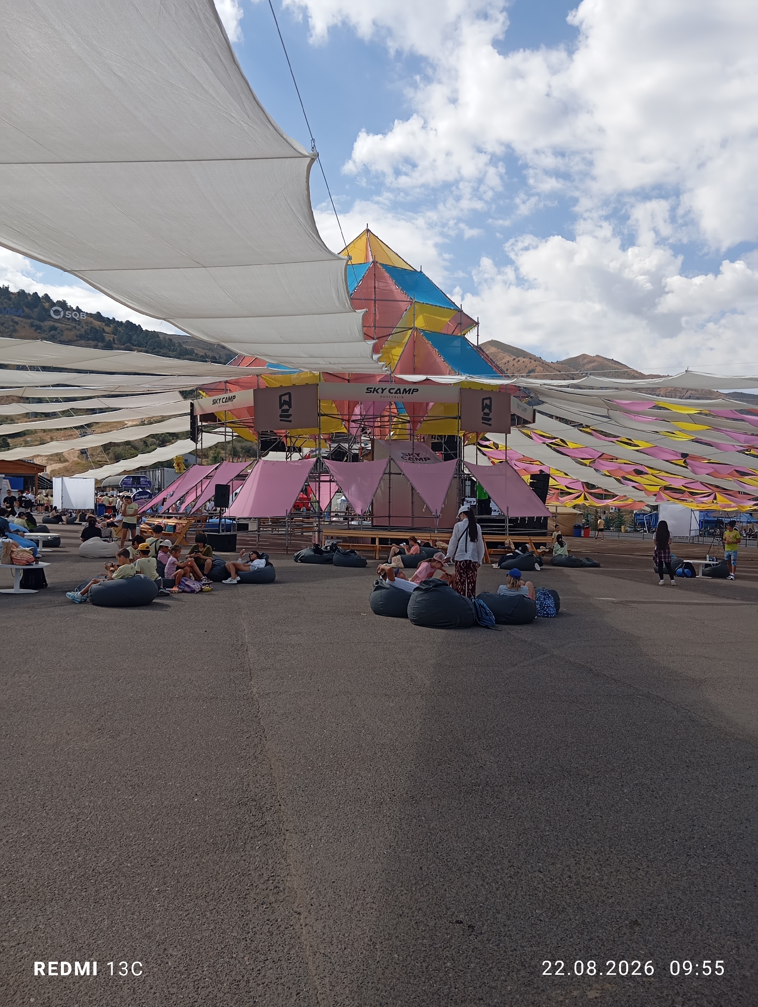 The image depicts an outdoor event scene at the ski resort in Amirsay, Uzbekistan. The foreground shows an asphalt ground with several people seated on large beanbag chairs, some of whom are interacting with each other. In the middle ground, a stage structure is visible, decorated with colorful triangular fabrics and signage that reads "SKY CAMP." There are also several pink tent-like structures set up. Beyond the stage, a large white canopy is stretched overhead, supported by poles, providing shade. In the background, hills and mountains are visible, indicating the mountainous terrain of the ski resort. The sky is partly cloudy. The overall impression is of a relaxed, possibly festival-like atmosphere.