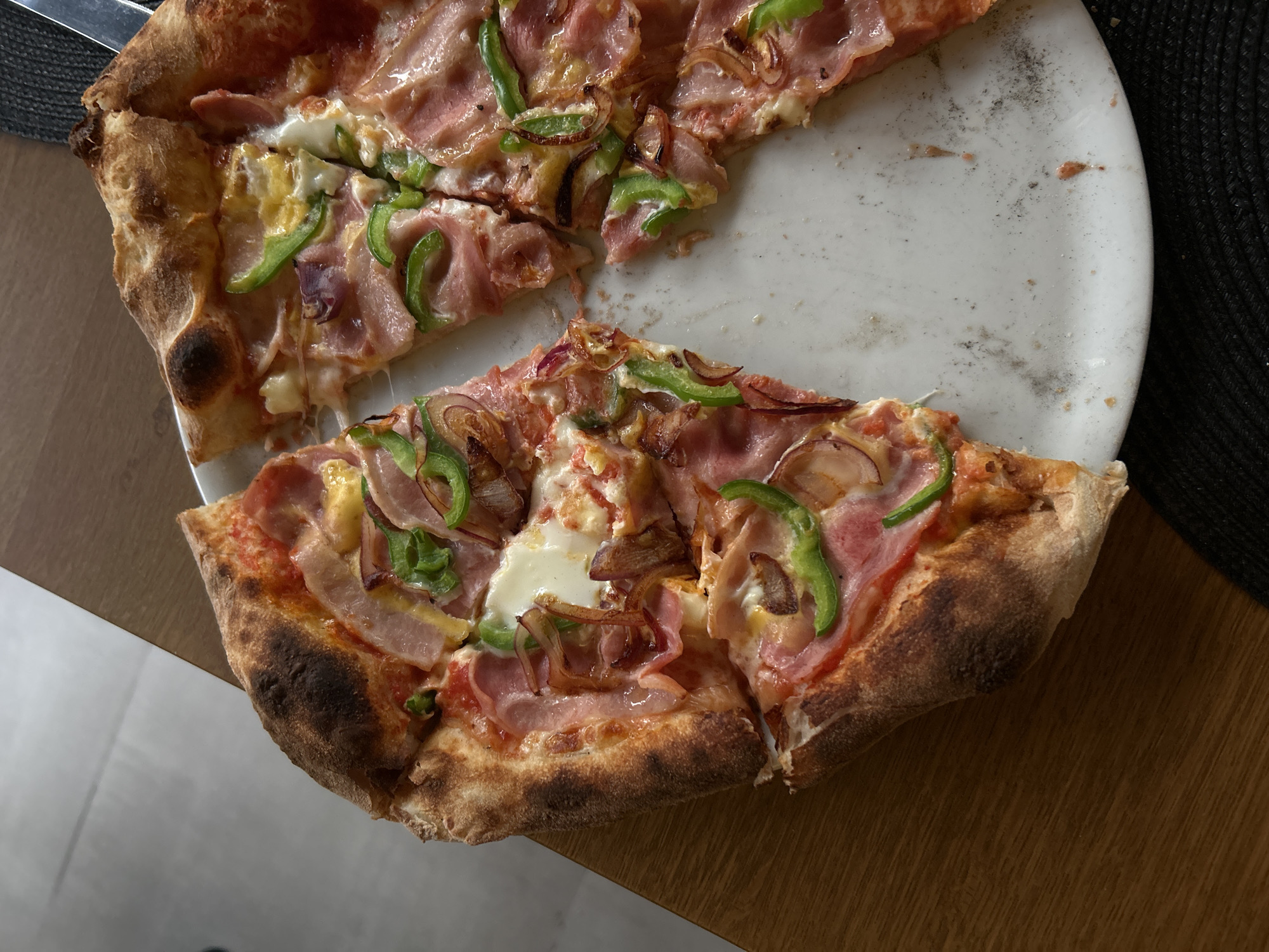 The image shows a partially eaten pizza on a white plate. The pizza has a thick, slightly charred crust and appears to be topped with ham, green peppers, and onions. The plate has some stains or food residue on it. The plate sits on a wooden surface. A knife rests beside the plate. A black placemat is visible in the background. It is not possible to determine the location of the image as Sofia, Bulgaria, based on the provided information.