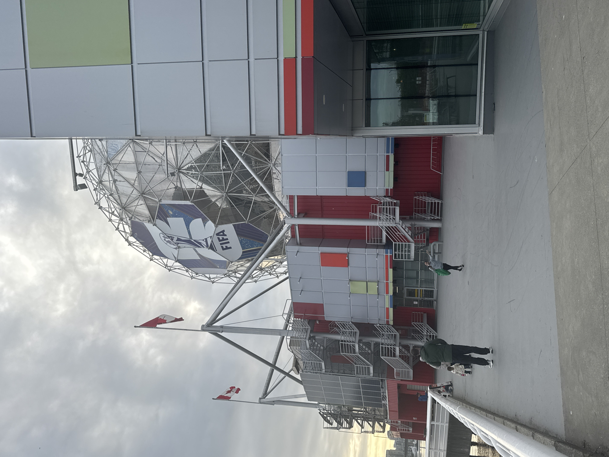 The image shows Science World, a geodesic dome in Vancouver, Canada, with a FIFA soccer ball design on its exterior. The structure is partially obscured by the corner of a building. Several people are walking near the base of the building. Canadian flags are visible on poles. The sky is overcast.
