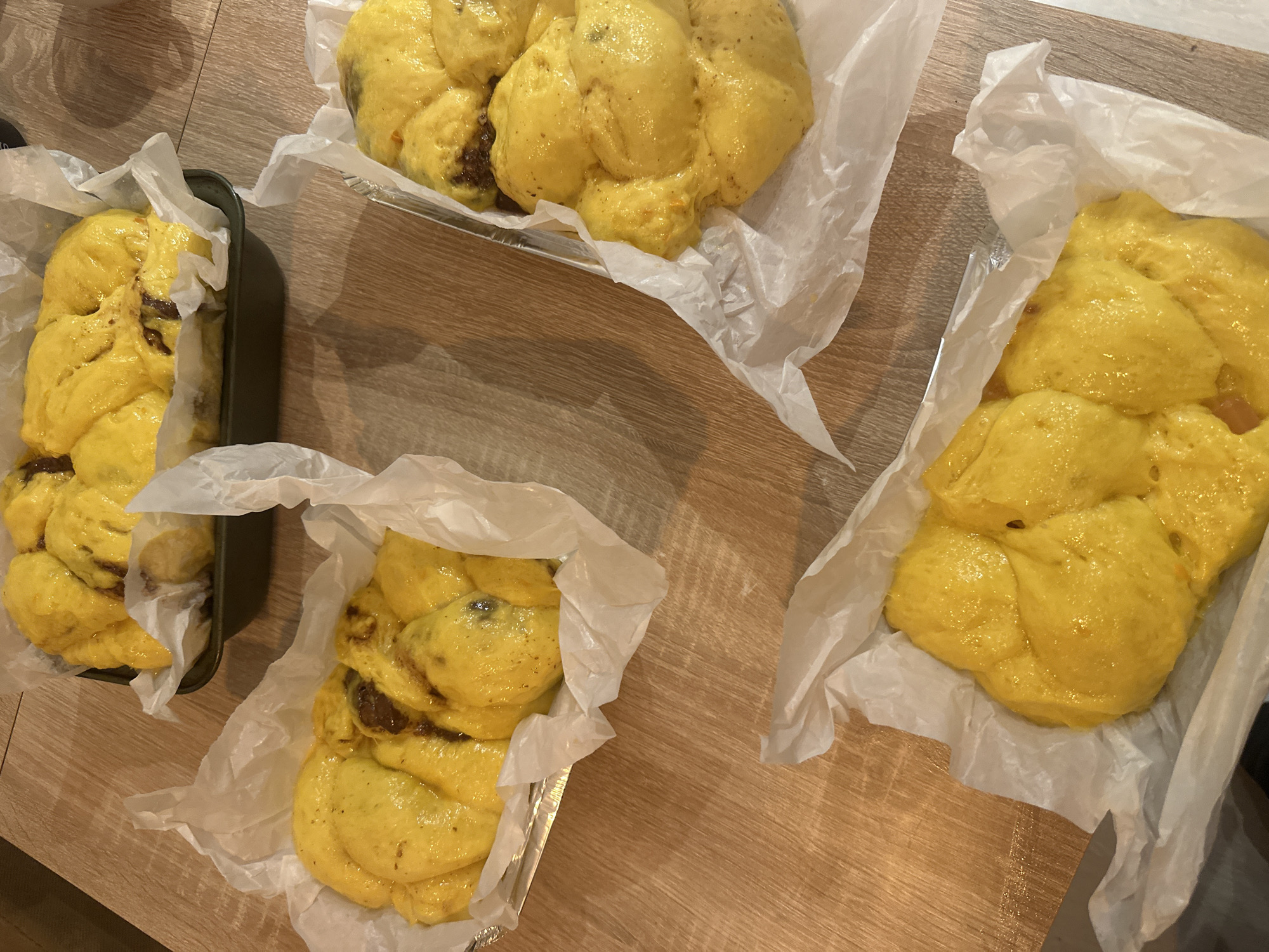 The image shows four unbaked yeast cakes on a light brown wooden table. Each cake sits in either a metal baking pan or on parchment paper. The cakes have a shiny yellow glaze and appear to have a chocolate filling. The scene is well-lit. No people are visible. The location is not discernible from the image.