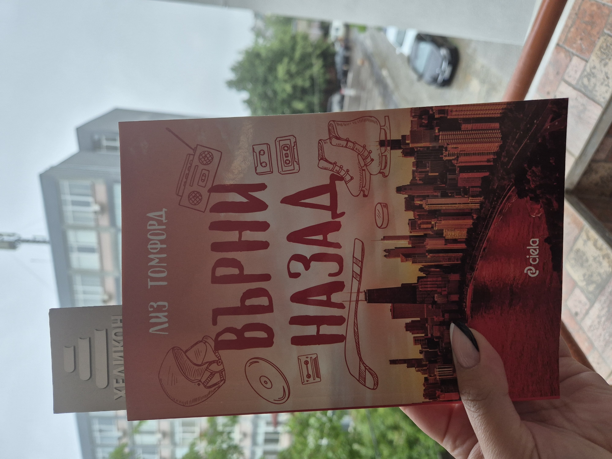 A person's hand, with a black-tipped French manicure, is holding a book outdoors in Sofia, Bulgaria. A gray bookmark, partially visible and inserted into the book, displays the name "МЕЛИКОН" along with a logo of three ascending vertical bars.
The book cover is prominently displayed, titled "ВЪРНИ НАЗАД" (meaning "Turn Back" or "Rewind") by author "ЛИЗ ТОМФОРД" (Liz Tomforde). The publisher's logo "@ciela" is also visible in the bottom right corner of the cover. The design features a vibrant, sunset-colored cityscape silhouette in shades of red, orange, and yellow, against which line art illustrations are superimposed. These illustrations include a boombox, two audio cassettes, a pair of ice skates, a hockey stick, an ice hockey puck, and a CD, suggesting themes of nostalgia and sports.
The background setting is an urban outdoor environment, possibly a balcony or terrace, evidenced by a tiled floor and a metal railing. Beyond, a modern building with multiple windows is visible, partially obscured by an out-of-focus gray sky. Blurred green trees and a dark car can also be seen, indicating a street or residential area. The lighting is soft and diffused, suggesting an overcast daytime.