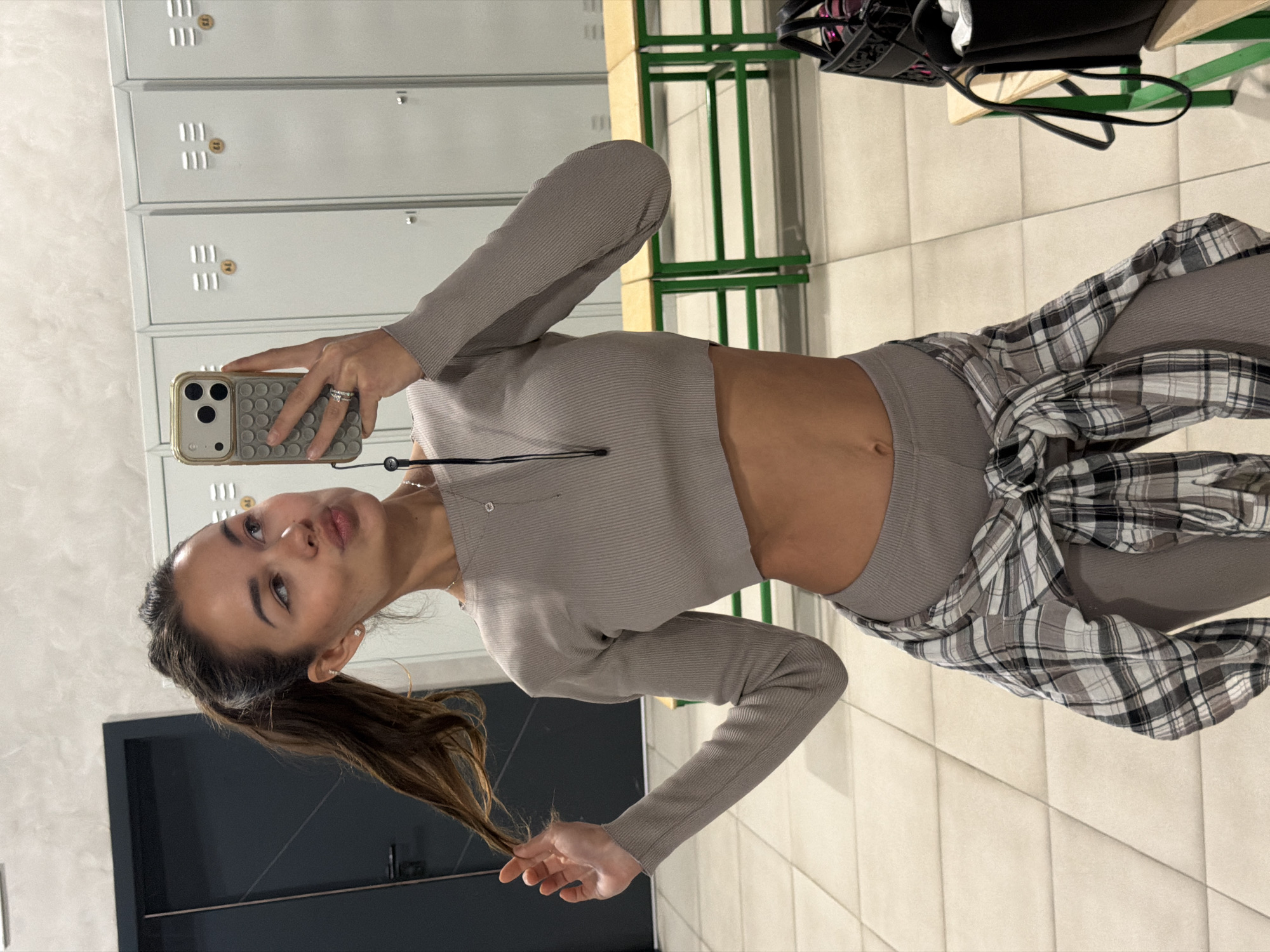 A young woman poses in a mirror selfie. She is wearing a gray, long-sleeved crop top, gray leggings, and a plaid shirt tied around her waist. She is holding a smartphone with a gray case. Her hair is pulled back in a ponytail. Lockers and a bench are visible in the background. The location could be a locker room or a gym, but there is no identifying information present to indicate the city or country.
